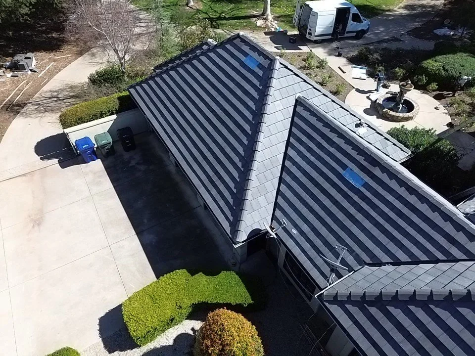 Aerial view of a house with a large driveway, a gray tiled roof, and a small garden with bushes and trees. A white van is parked on the street near the house. There is also a small fountain or pond in the backyard.