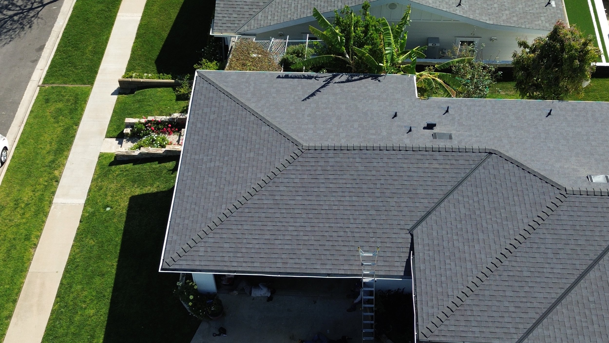 An aerial view of a house with a gray roof, surrounded by a sidewalk, green lawns, trees, and neighboring houses with similar roofs.