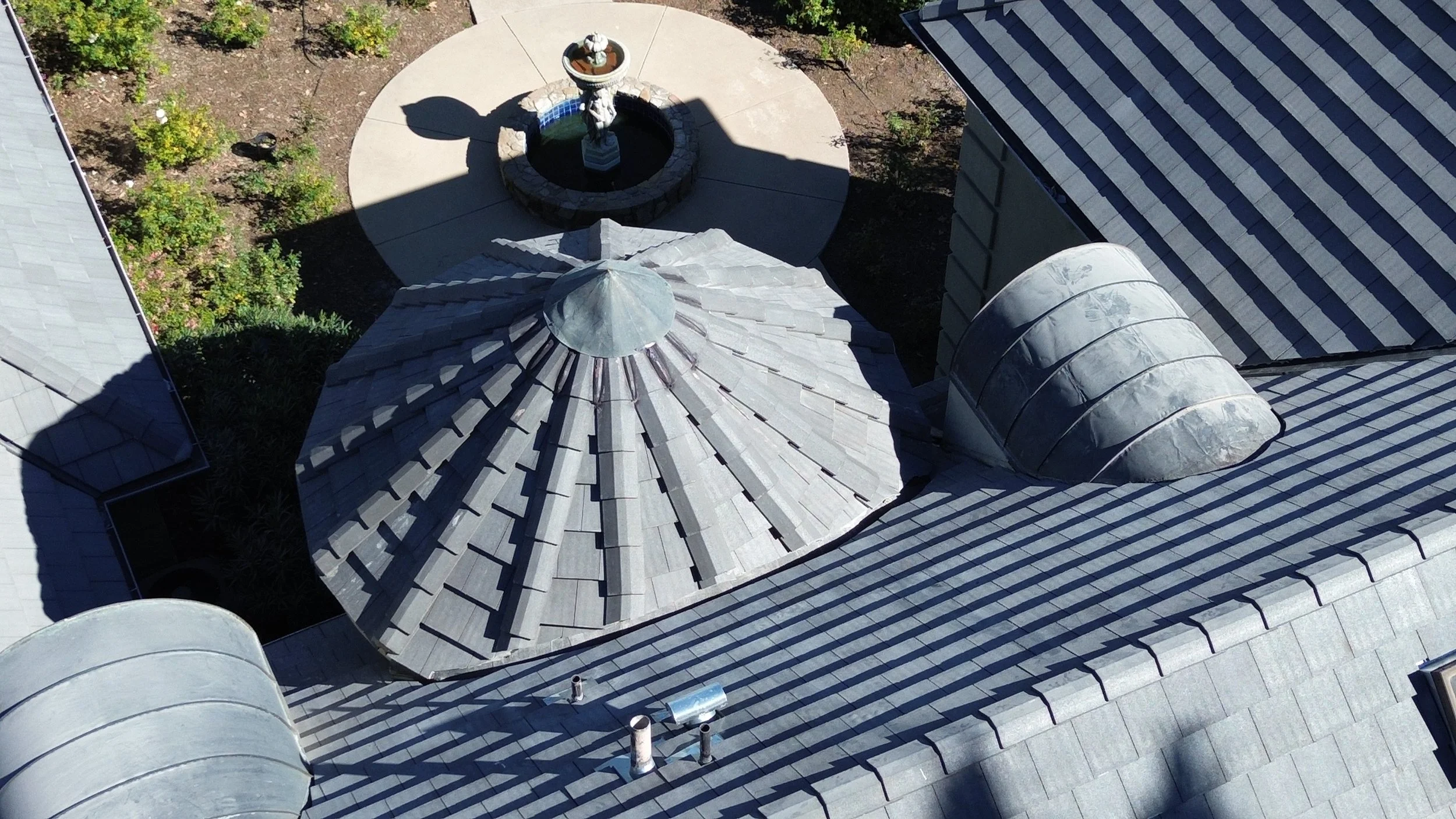 An aerial view of rooftops with gray shingles, featuring a small dome-shaped structure and chimneys. In the background, there is a garden area with a circular patio, a fountain, and some greenery.