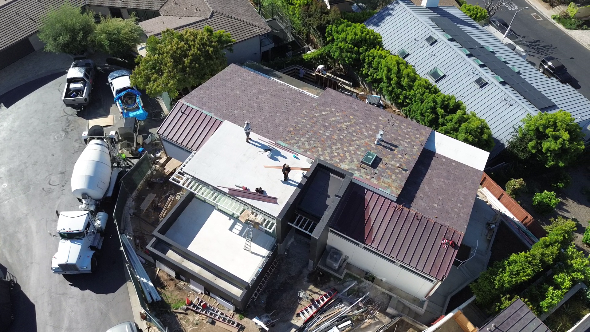 Aerial view of a construction site on a residential roof with workers, building materials, and vehicles, including trucks and construction equipment, surrounding the house.