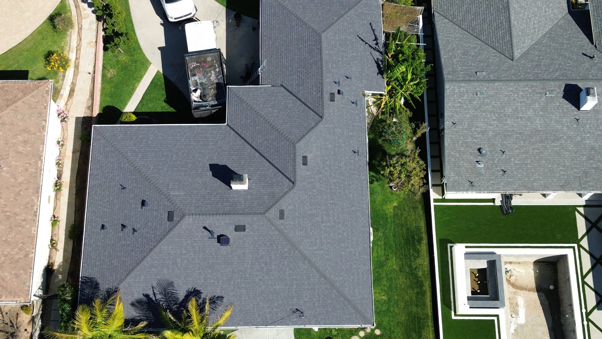 Aerial view of residential houses with gray shingle roofs, green lawns, trees, and a backyard with patio furniture.