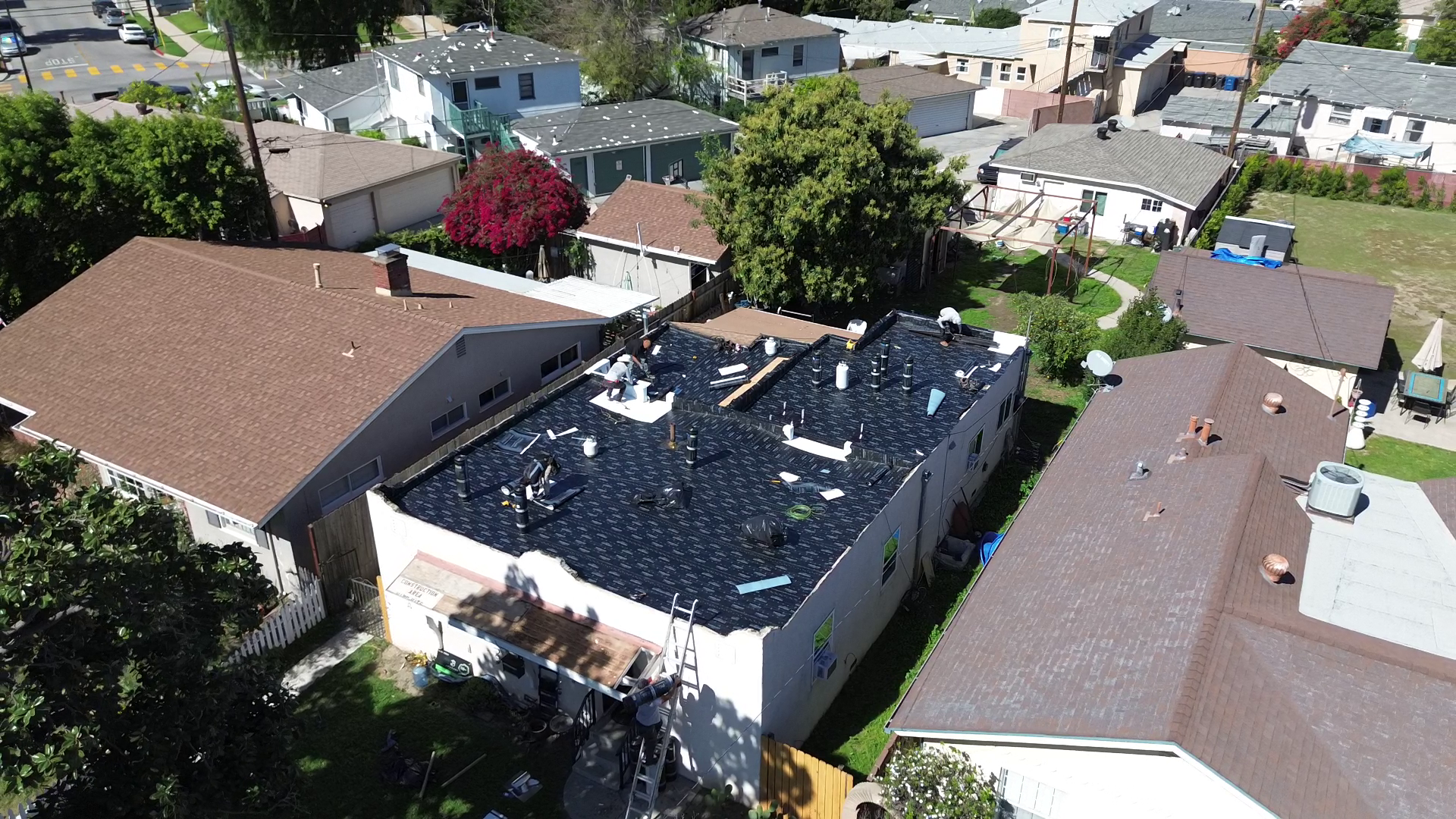 Aerial view of a residential neighborhood showing houses with brown, gray, and blue roofs. One house's roof is under construction with workers installing a black roofing material. Surrounding houses have trees, lawns, and outdoor furniture.
