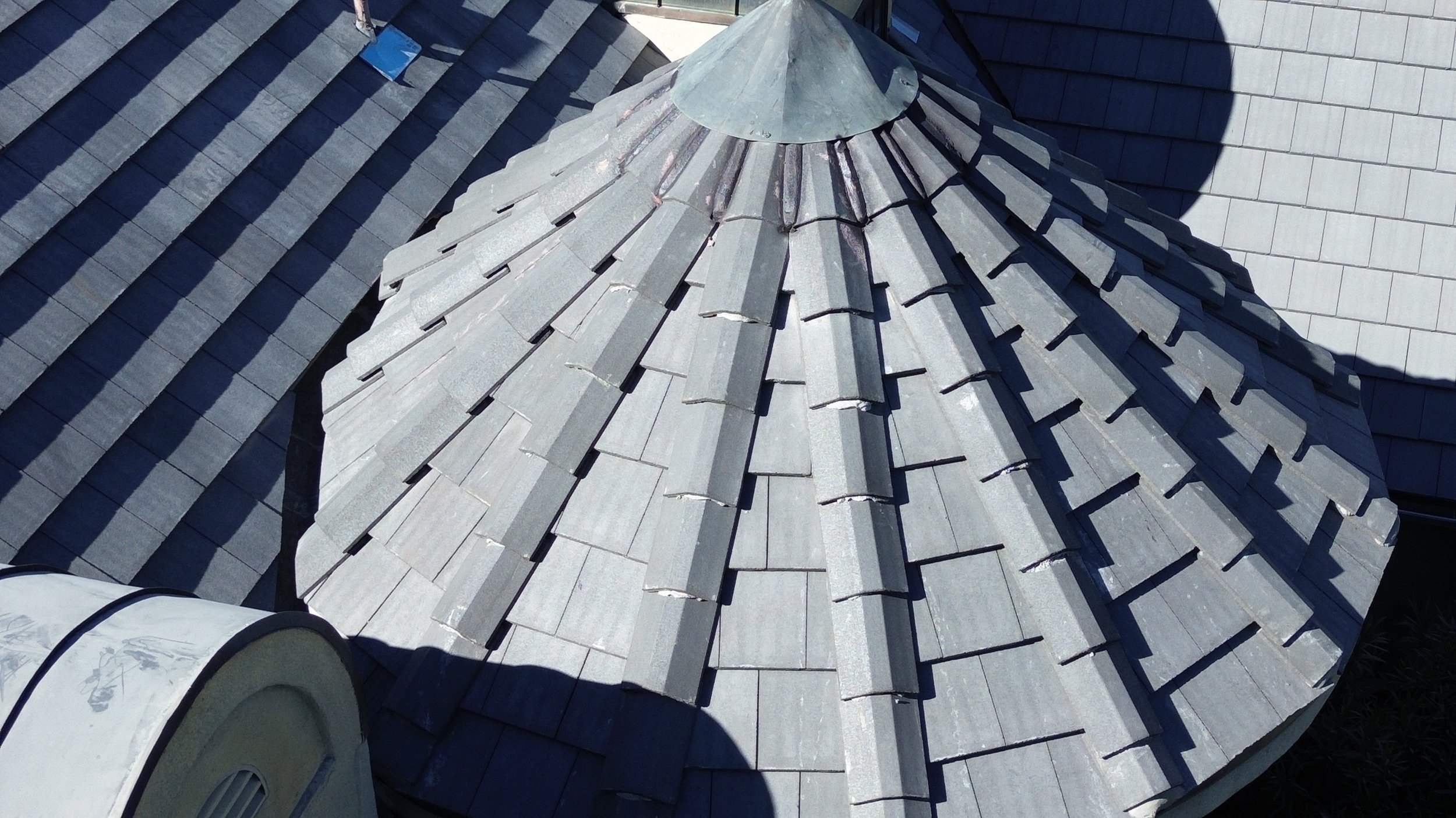 Close-up view of a gray, shingle-style roof with a conical vent cap at the peak, surrounded by other sections of the roof.