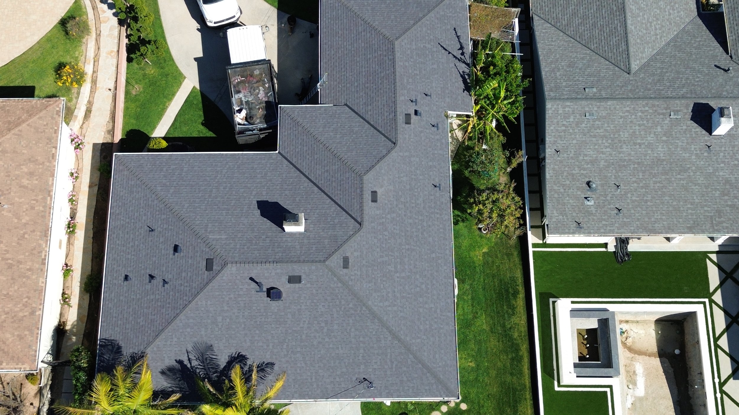 Aerial view of residential houses with grey and brown shingle roofs, green lawns, a white fence, trees, and a backyard with patio and landscaping.