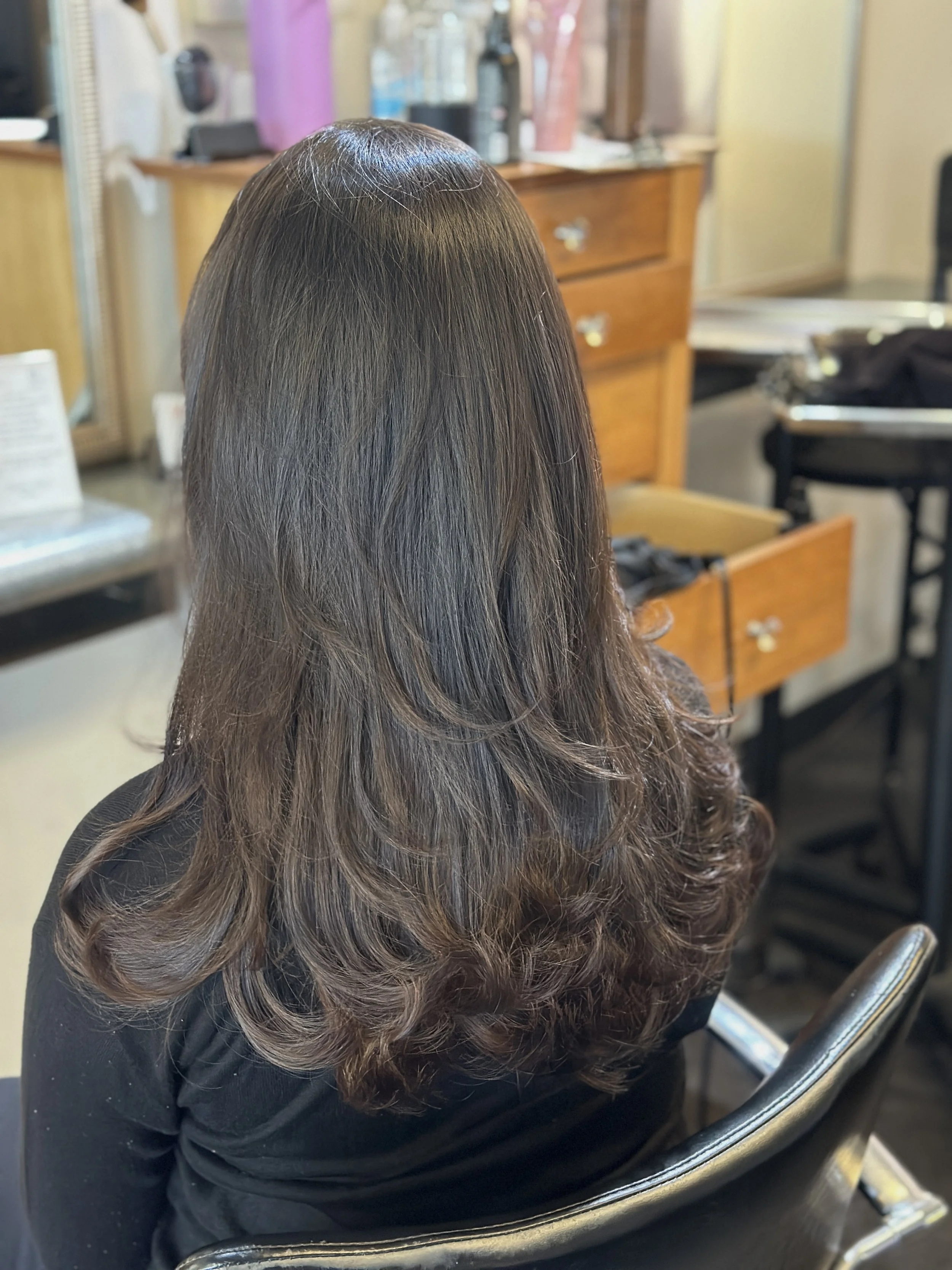 Woman with long, wavy, dark brown hair sitting in a salon chair, with hair styled and shiny, in a salon setting.