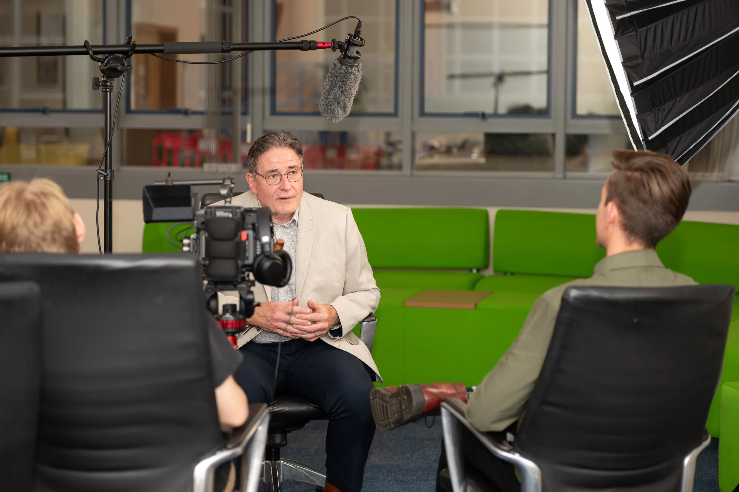 An interview scene with a man in a beige blazer being recorded by a camera crew, including a camera operator and sound technician, in an indoor setting with large windows and green seating.