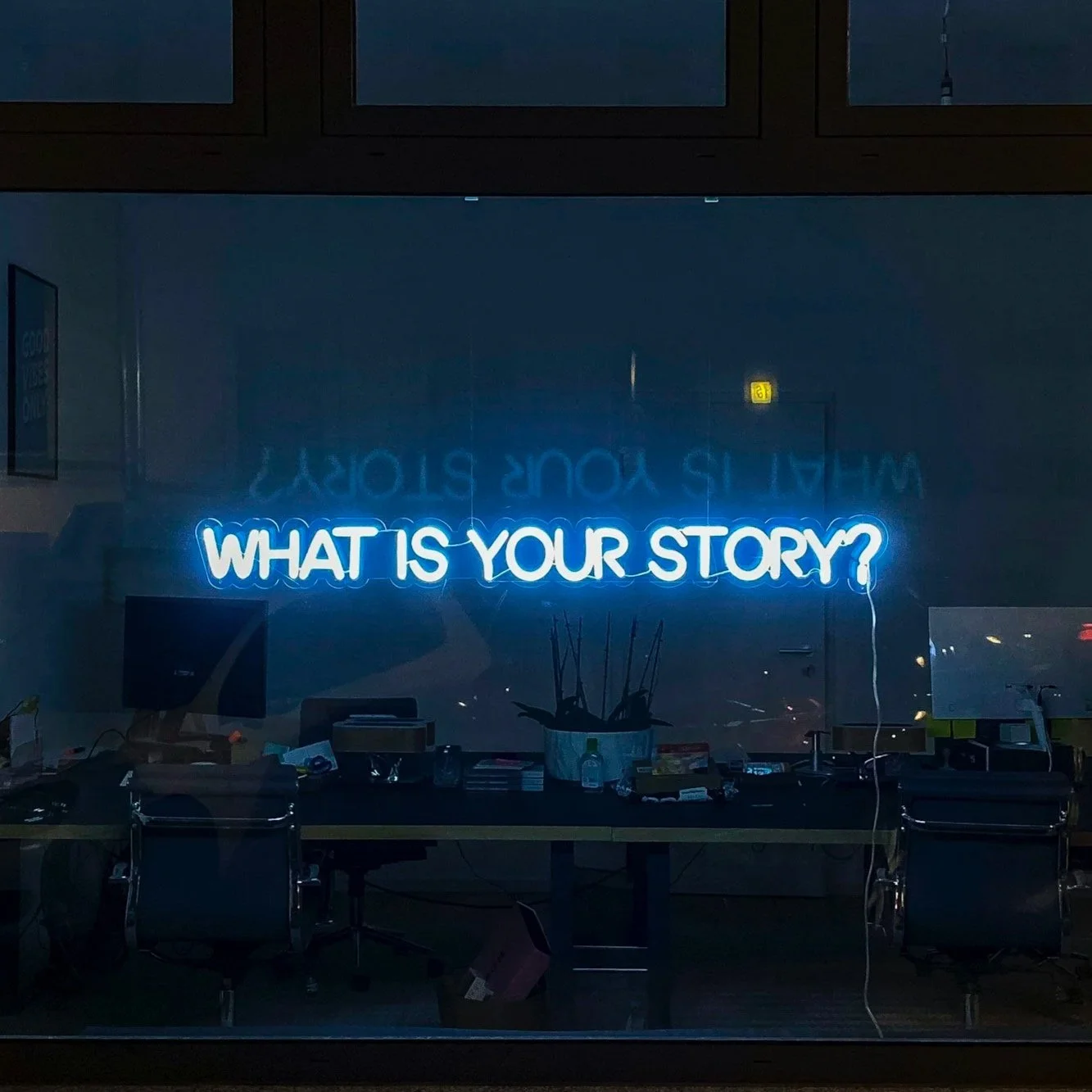 Neon sign reading 'What is your story?' in a dark office space with desks, chairs, and office supplies visible through a window.