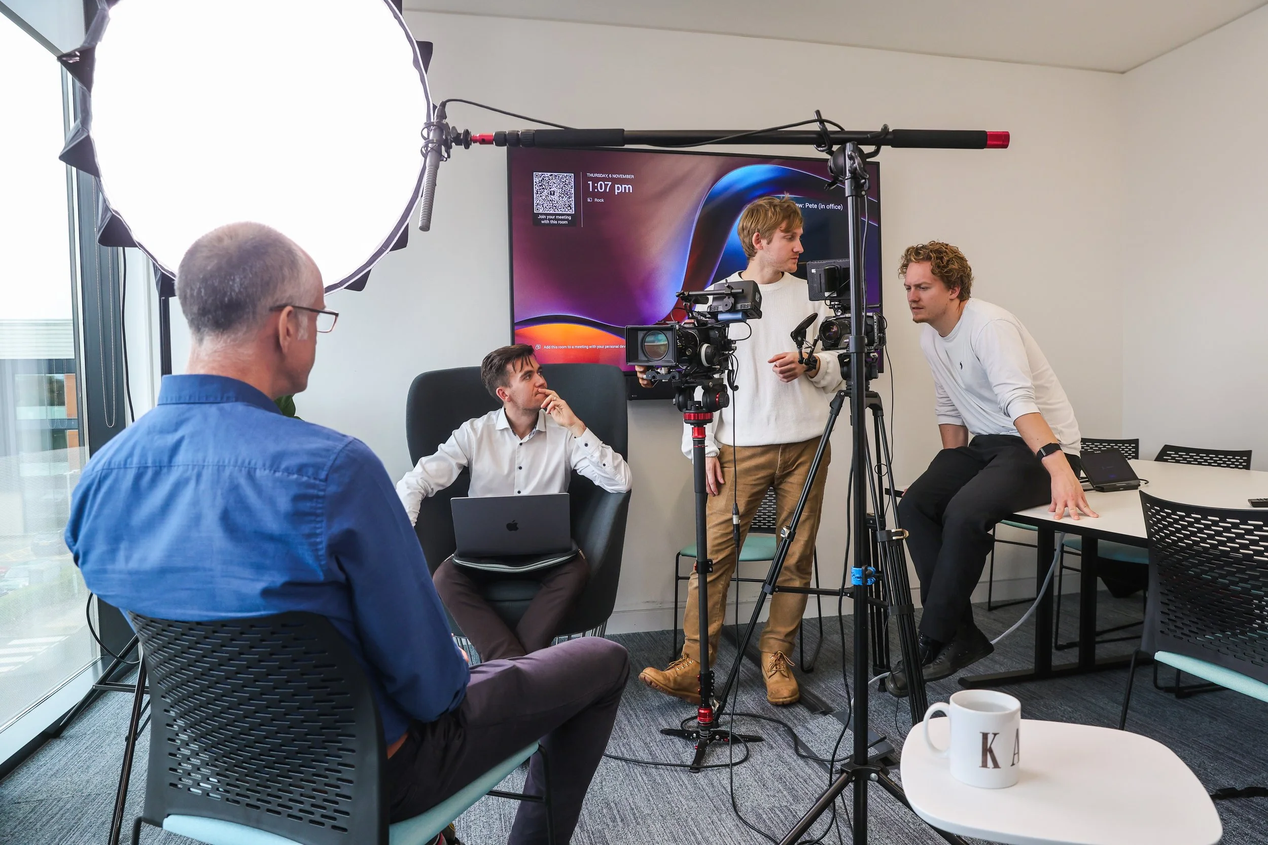 A behind-the-scenes shot of a video or photo shoot in an office setting with five men working with cameras, a large softbox light, and a computer monitor on the wall.