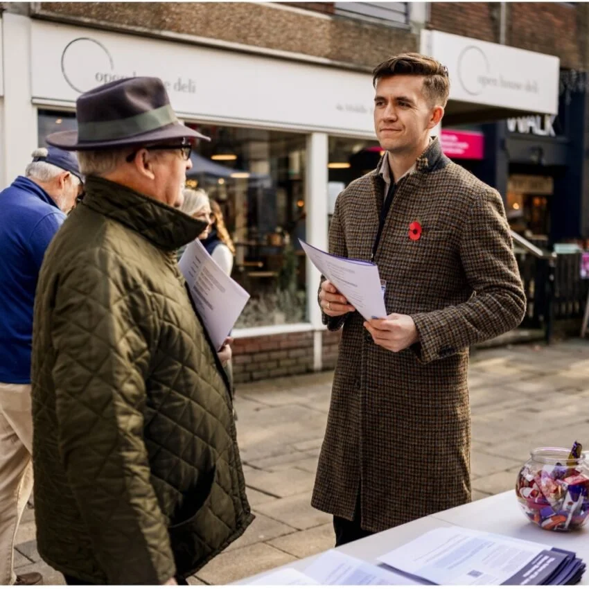 Two men having a conversation outdoors, holding papers, with a table and jar of candies nearby on a city sidewalk.