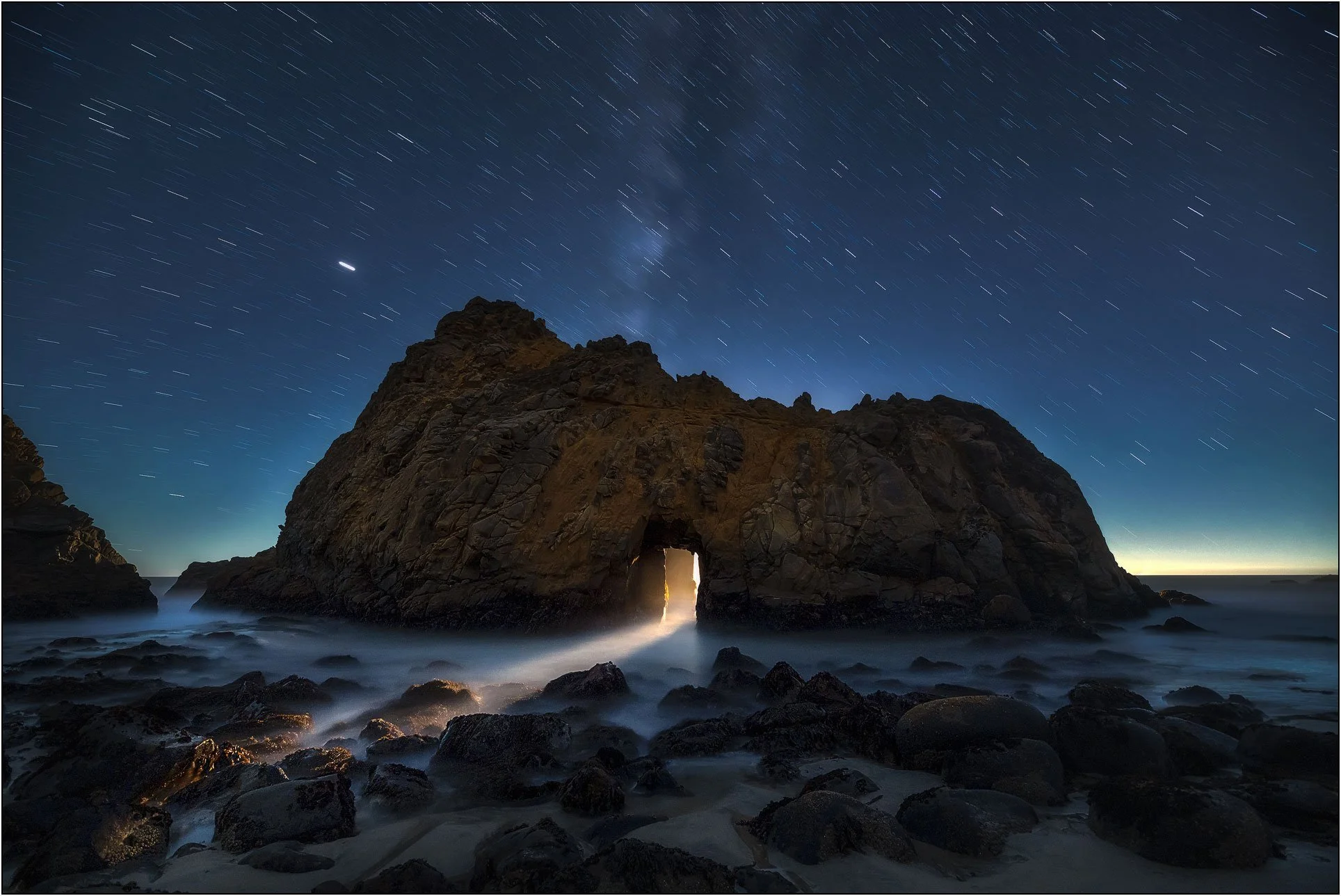Moonlit sea arch with light shining through under a starry night sky