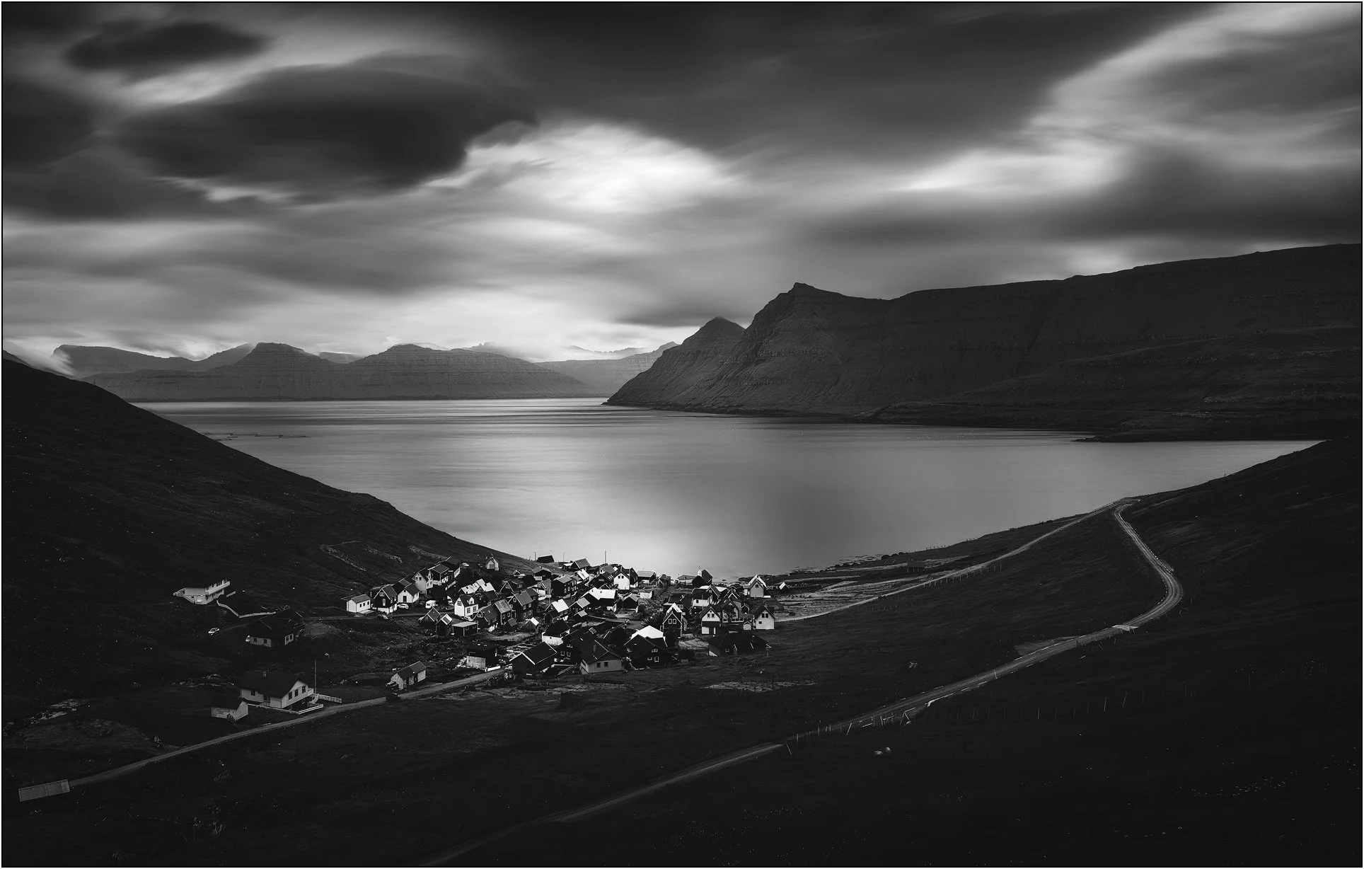 Black and white landscape of Funningur village in the Faroe Islands, with a fjord, mountains, and long exposure clouds and water.