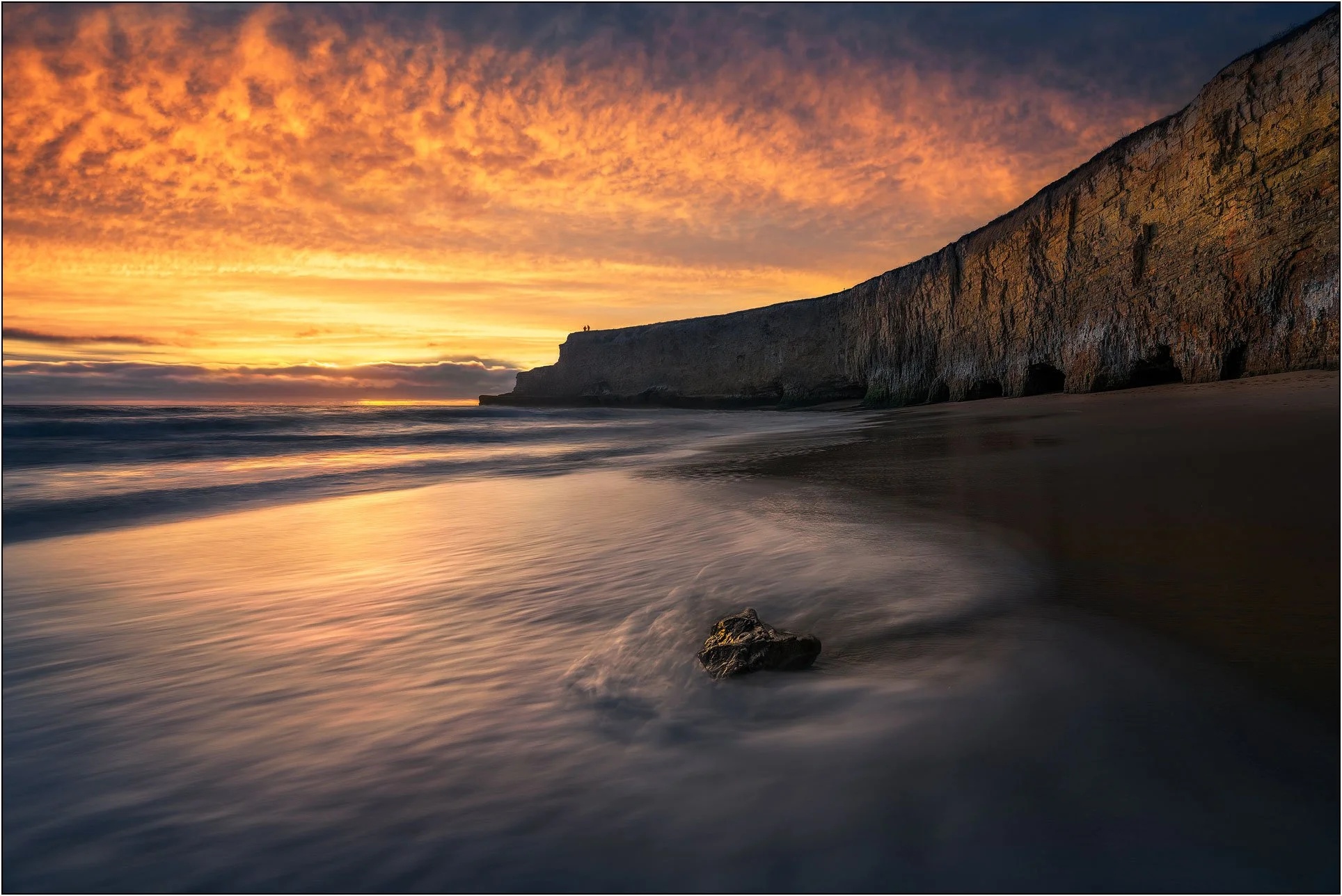 Seaside cliff at sunset with glowing orange clouds, gentle waves, and a single rock in the foreground