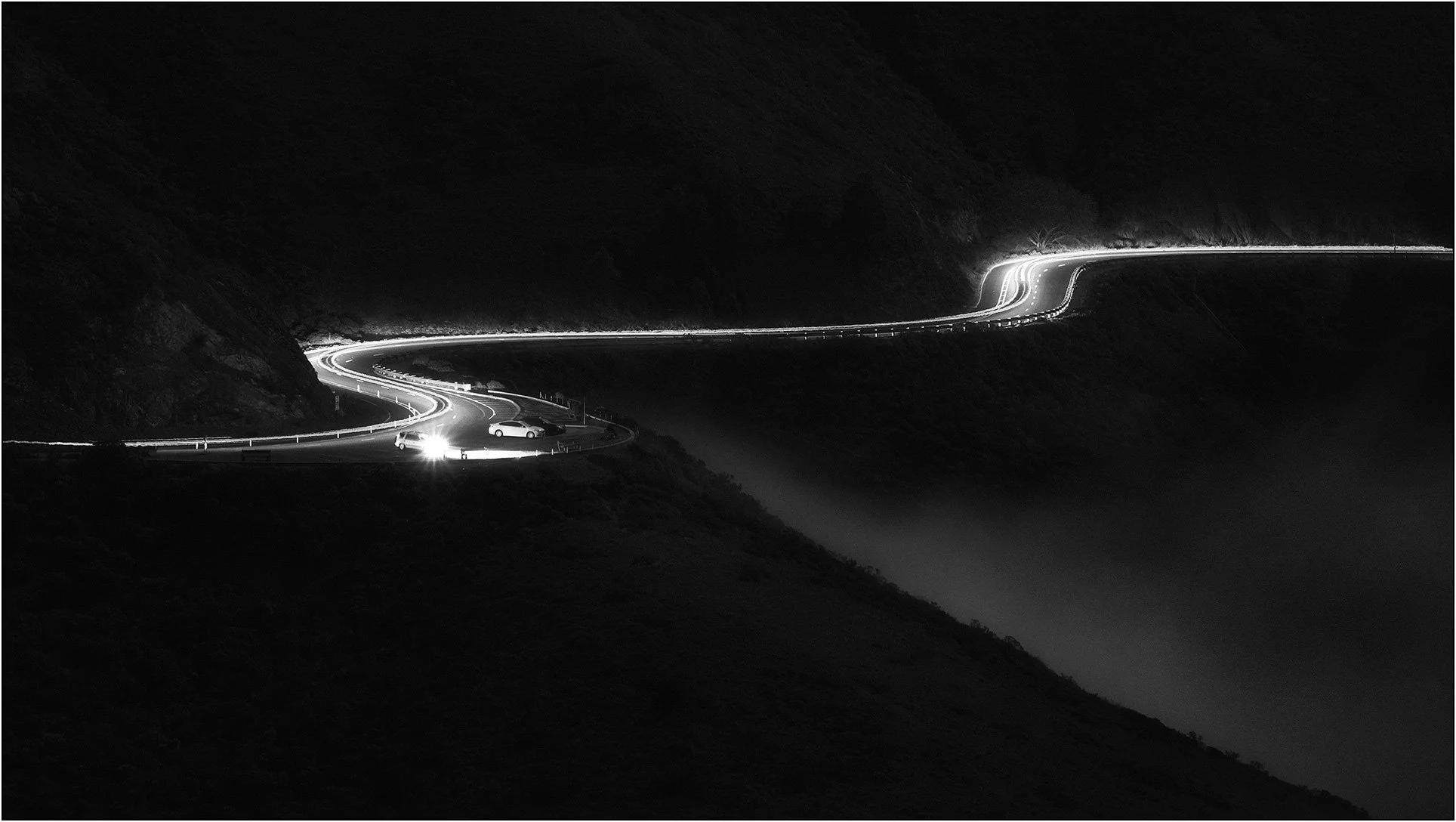 Winding coastal road at night captured with long exposure light trails against a dark hillside