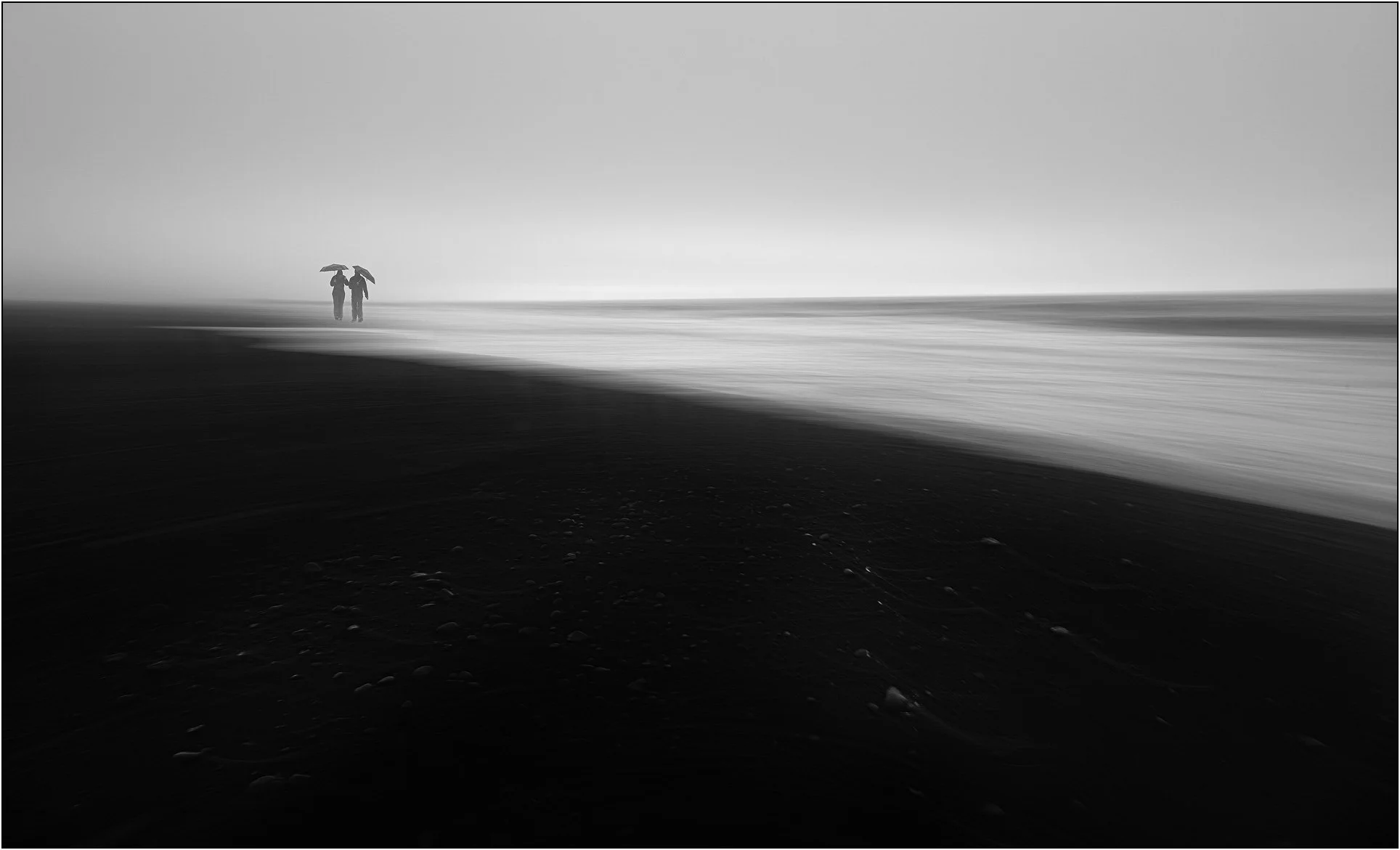 Black and white image of two people with umbrellas walking on the black beach in Iceland, waves softened by long exposure.