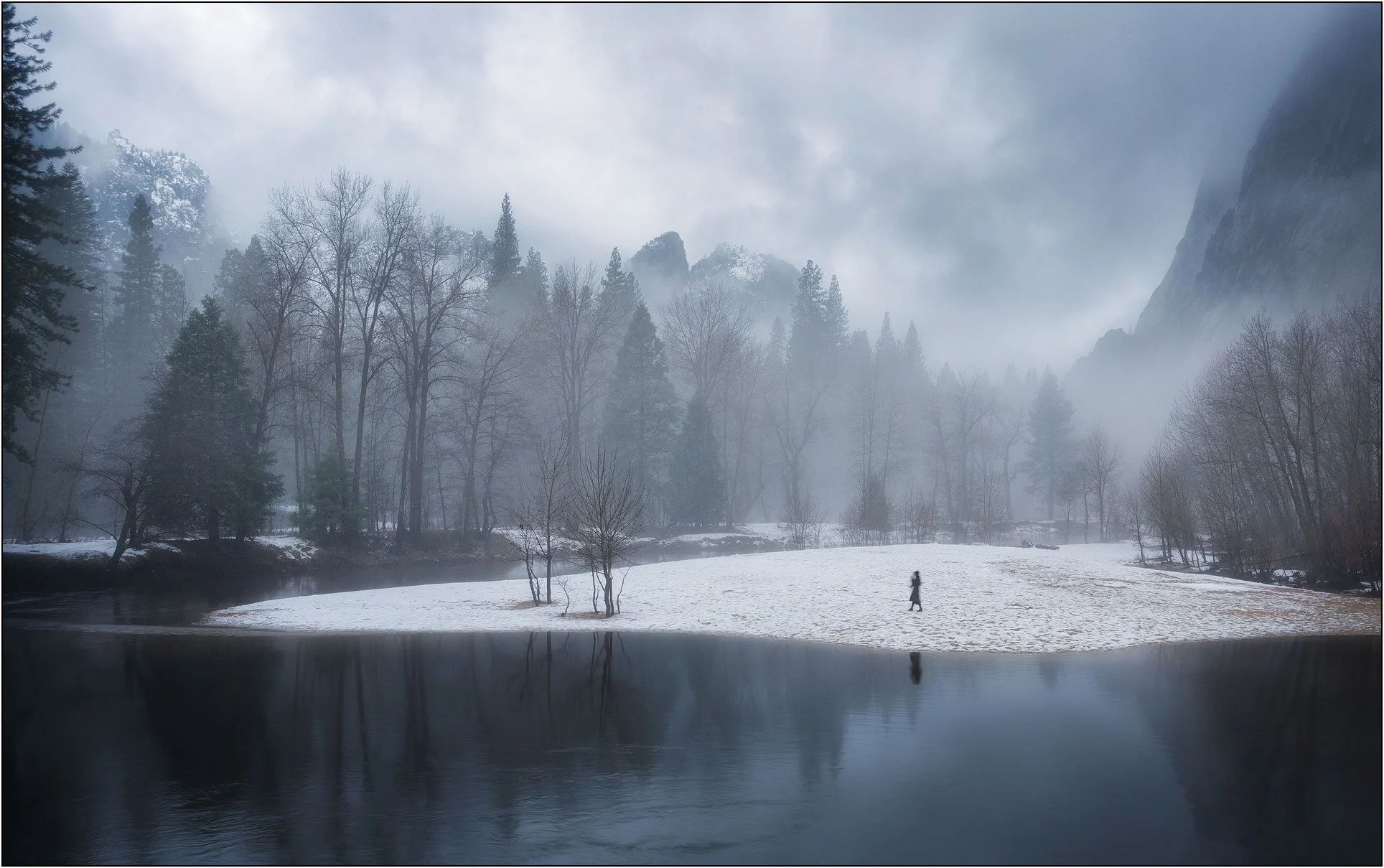 A lone figure walking across a snowy riverbank in a misty forest landscape of Yosemite valley