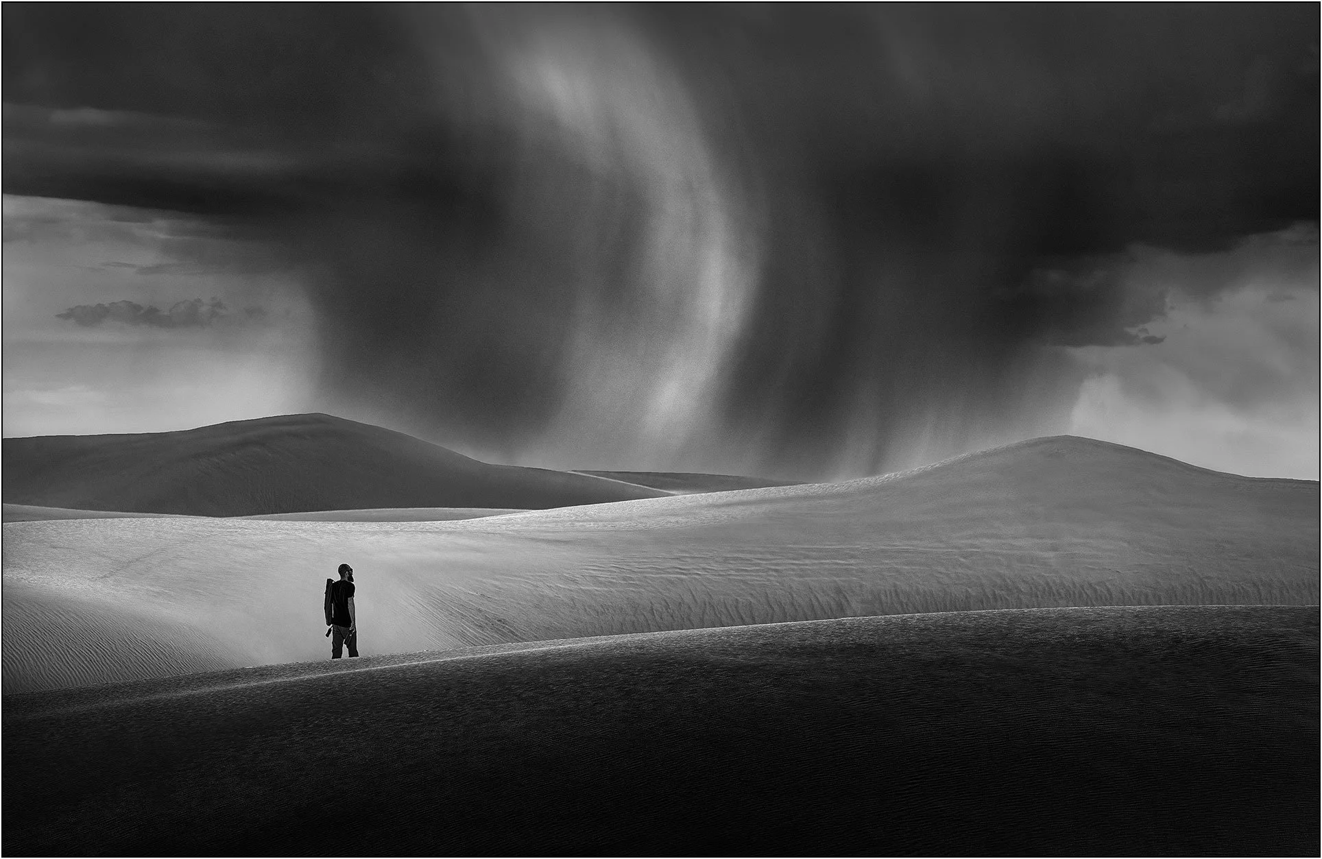 A person standing on sand dunes under dramatic swirling clouds in black and white