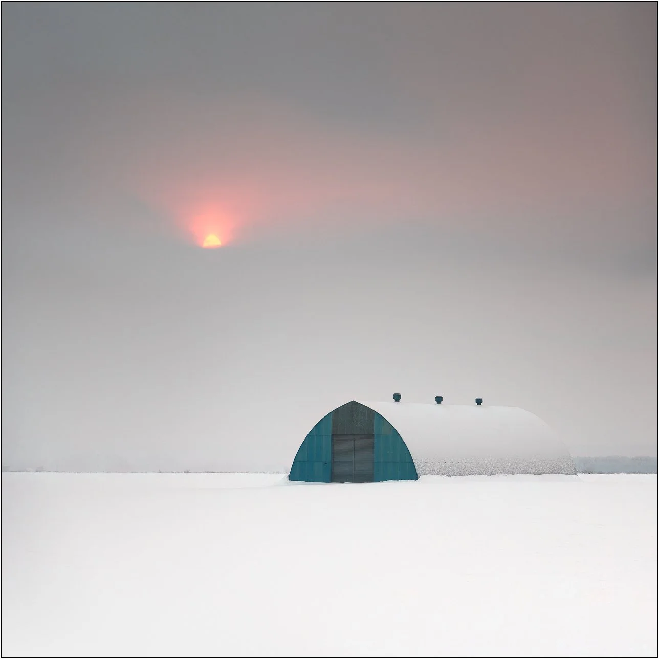 A lone barn in a snow-covered field under a soft sunrise partially obscured by mist in Hokkaido, Japan.