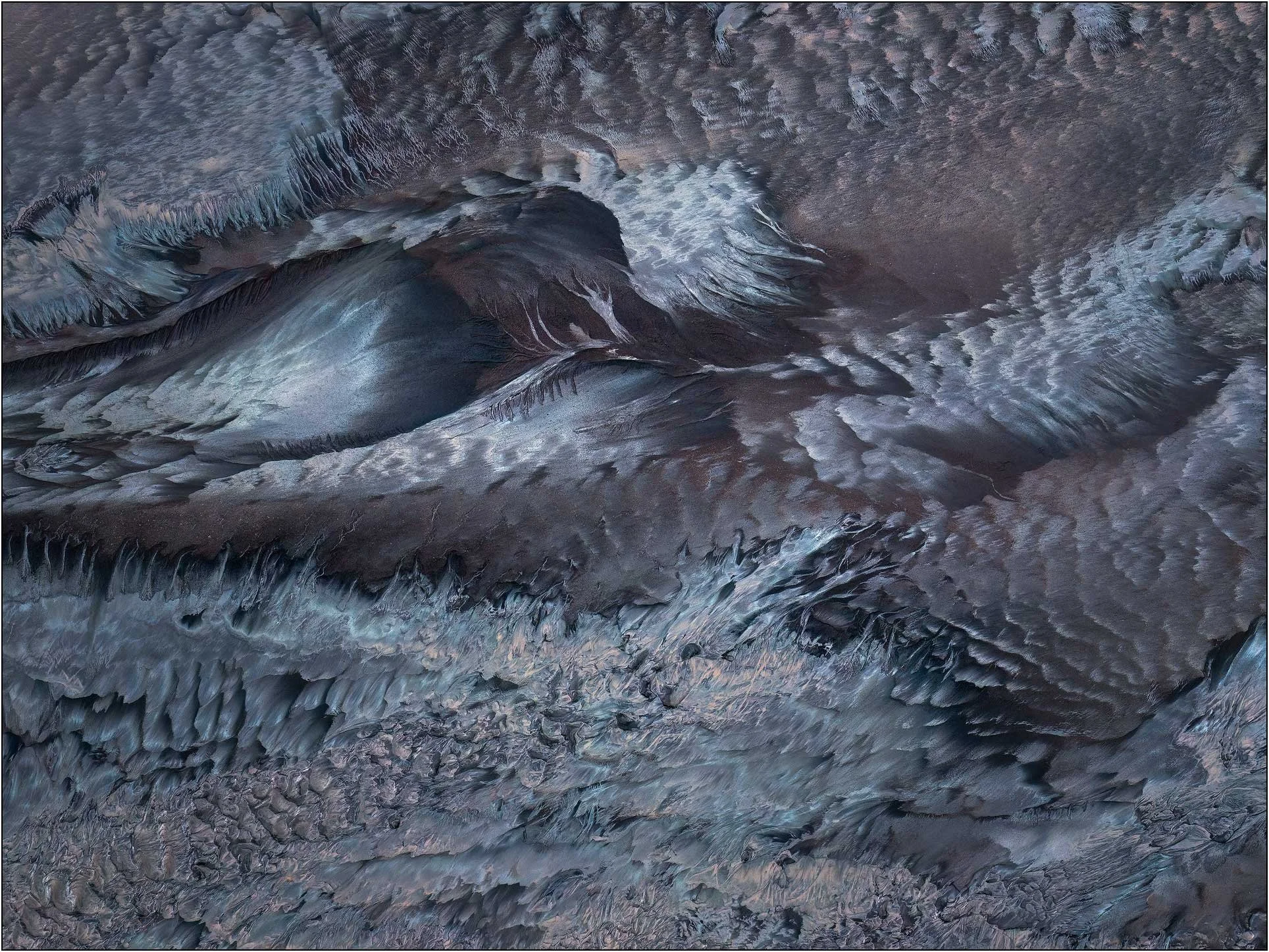 Aerial photography of the Qiantang River tidal flats in China, where tidal erosion shapes layered sediment into dramatic ridges, ripples, and flowing textures. Cool blue-gray tones and organic patterns transform the exposed riverbed into an abstract 
