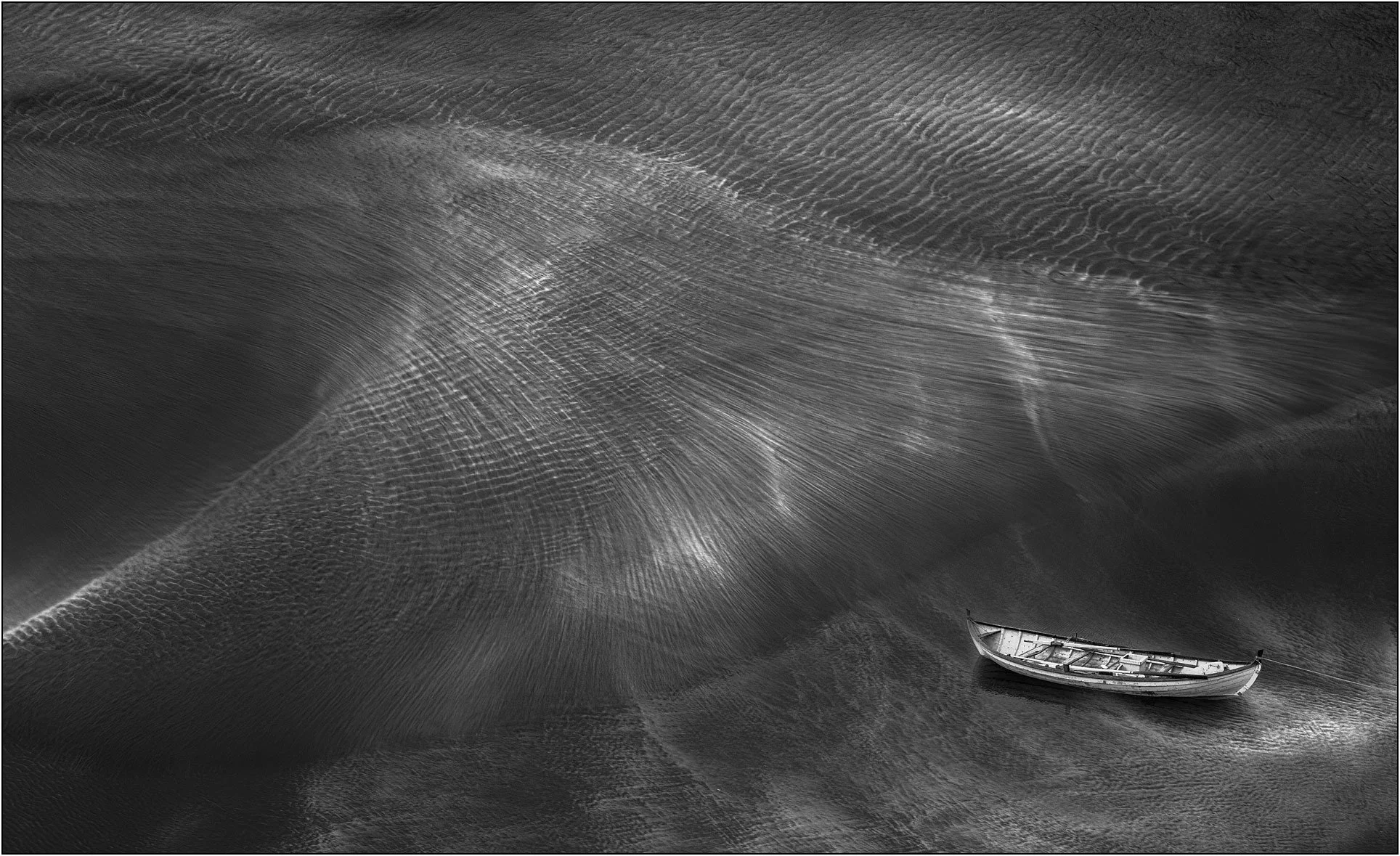 Black and white image of a boat on a beach with waves sweep in, creating textures of water and beach.