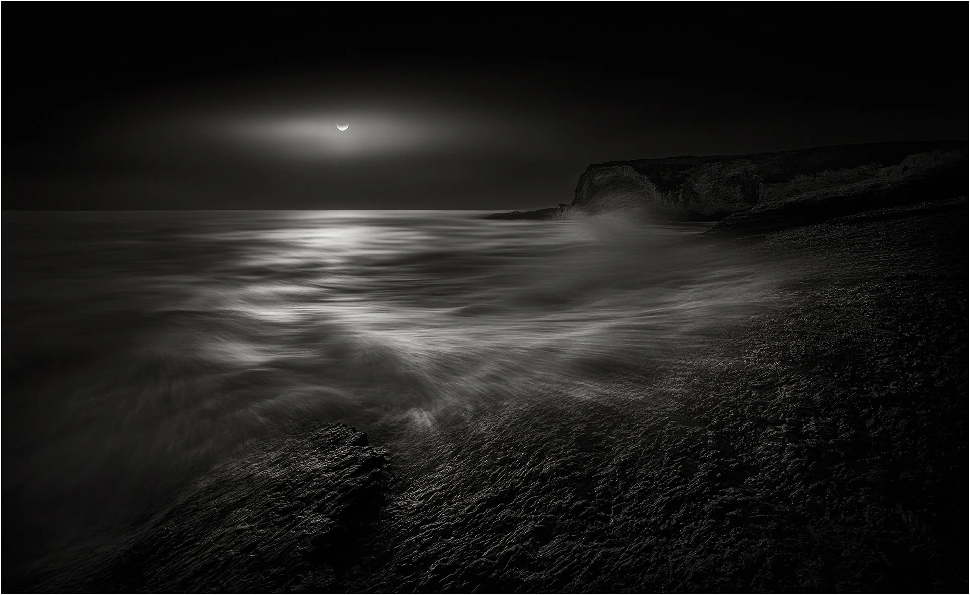 Black and white night seascape with moonlight reflecting on ocean waves and a distant cliff