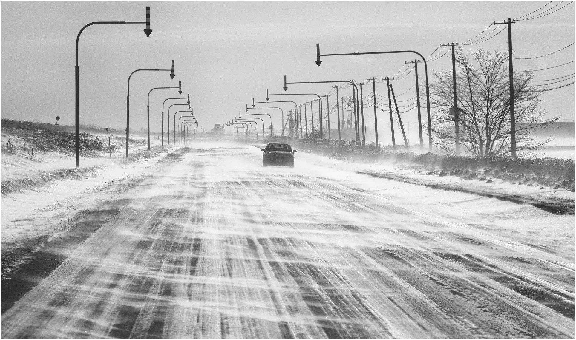 A car driving along a snowy road shaped by strong wind patterns in Hokkaido, Japan.