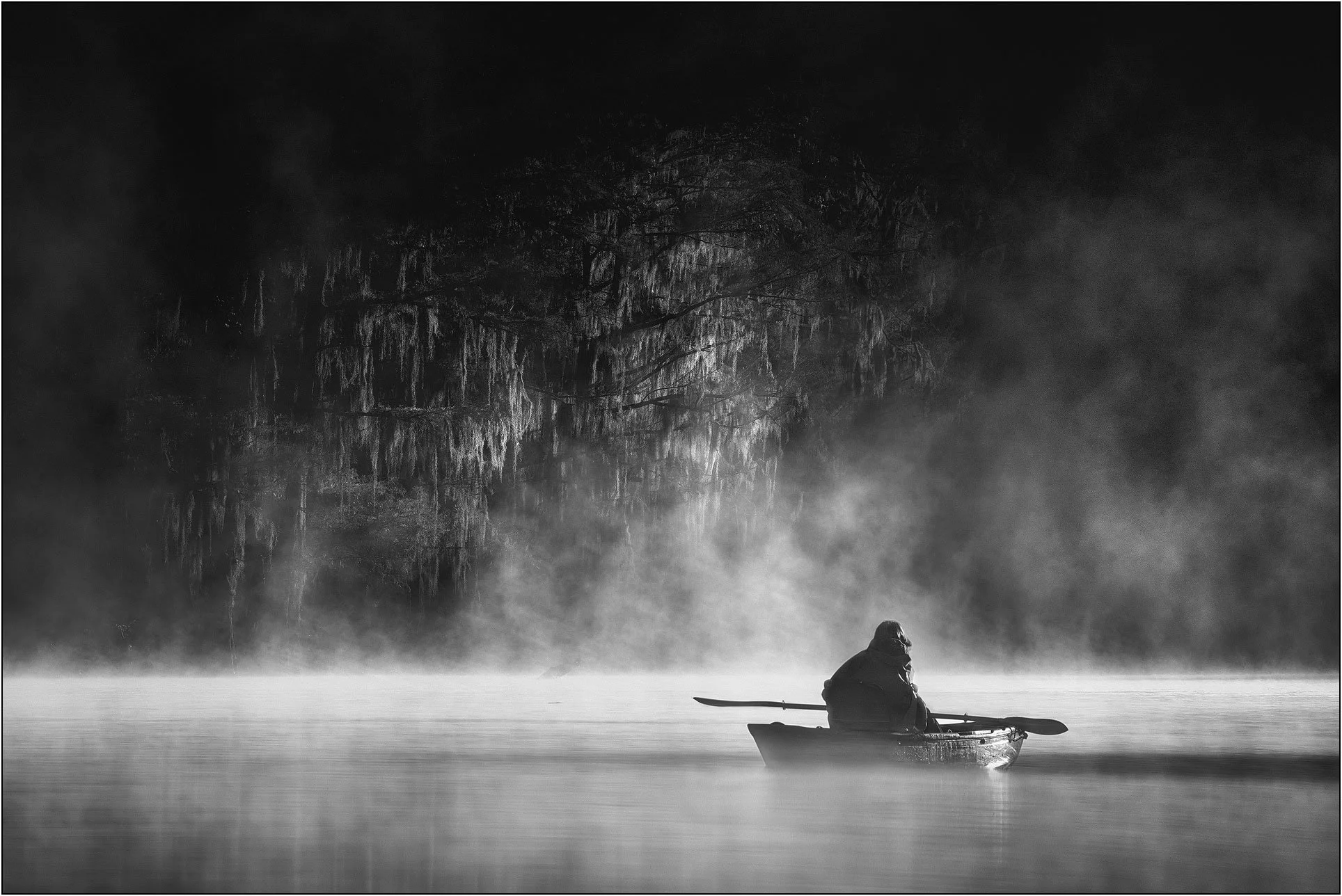 Black and white photograph of a woman kayaking in misty southern wetlands with bald cypress trees under morning light.