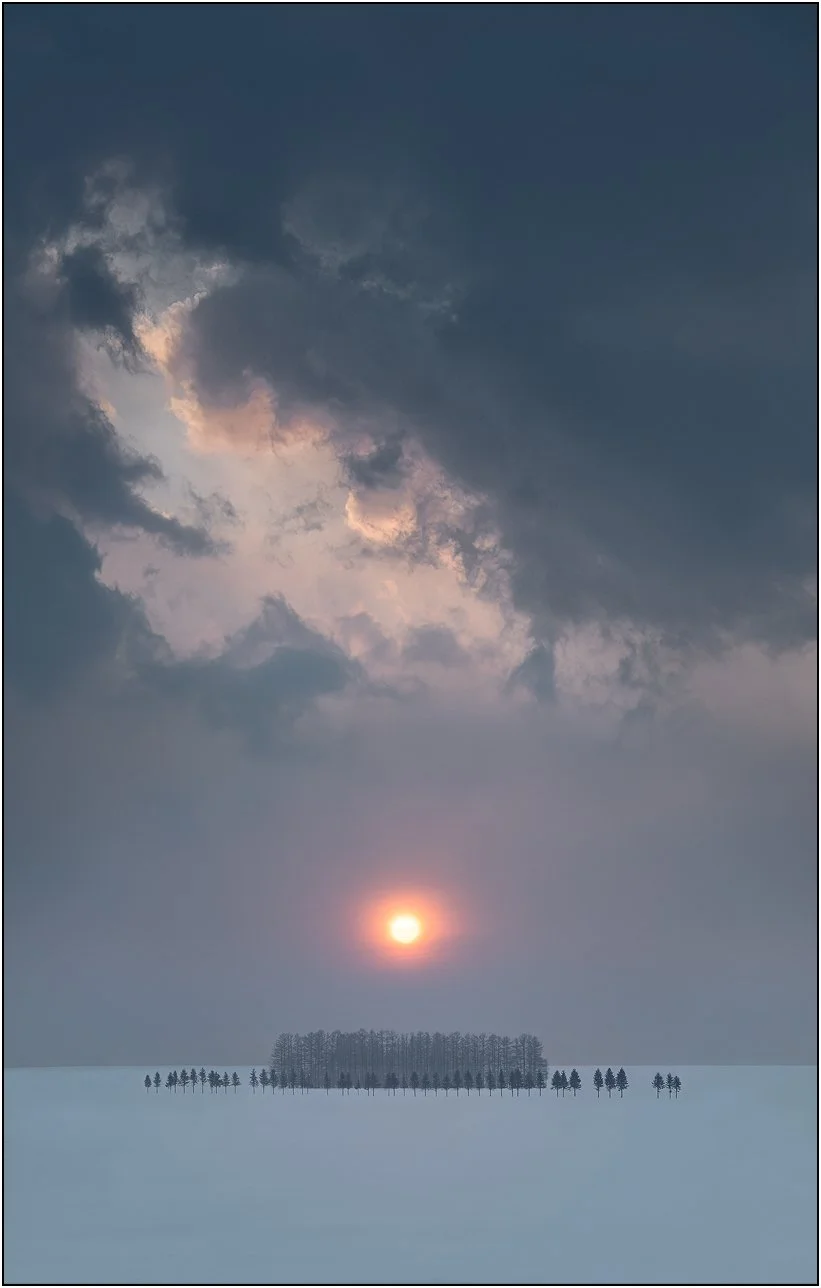 Snow-covered trees beneath a glowing sunrise with dramatic clouds above in Hokkaido, Japan.