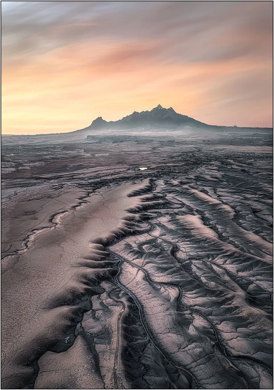 Eroded badlands leading toward a distant mountain under a soft pastel sky