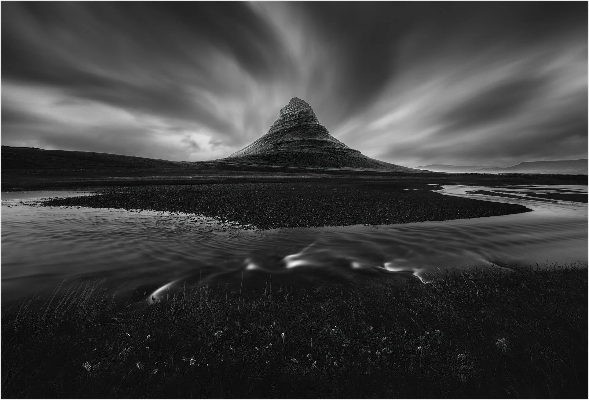 Black and white long exposure image of Kirkjufell, Iceland, with grass and water flow in the foreground and mountain in the background.