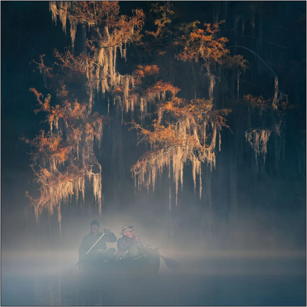 Two men kayaking in misty southern wetlands with morning light on bald cypress trees in fall colors.