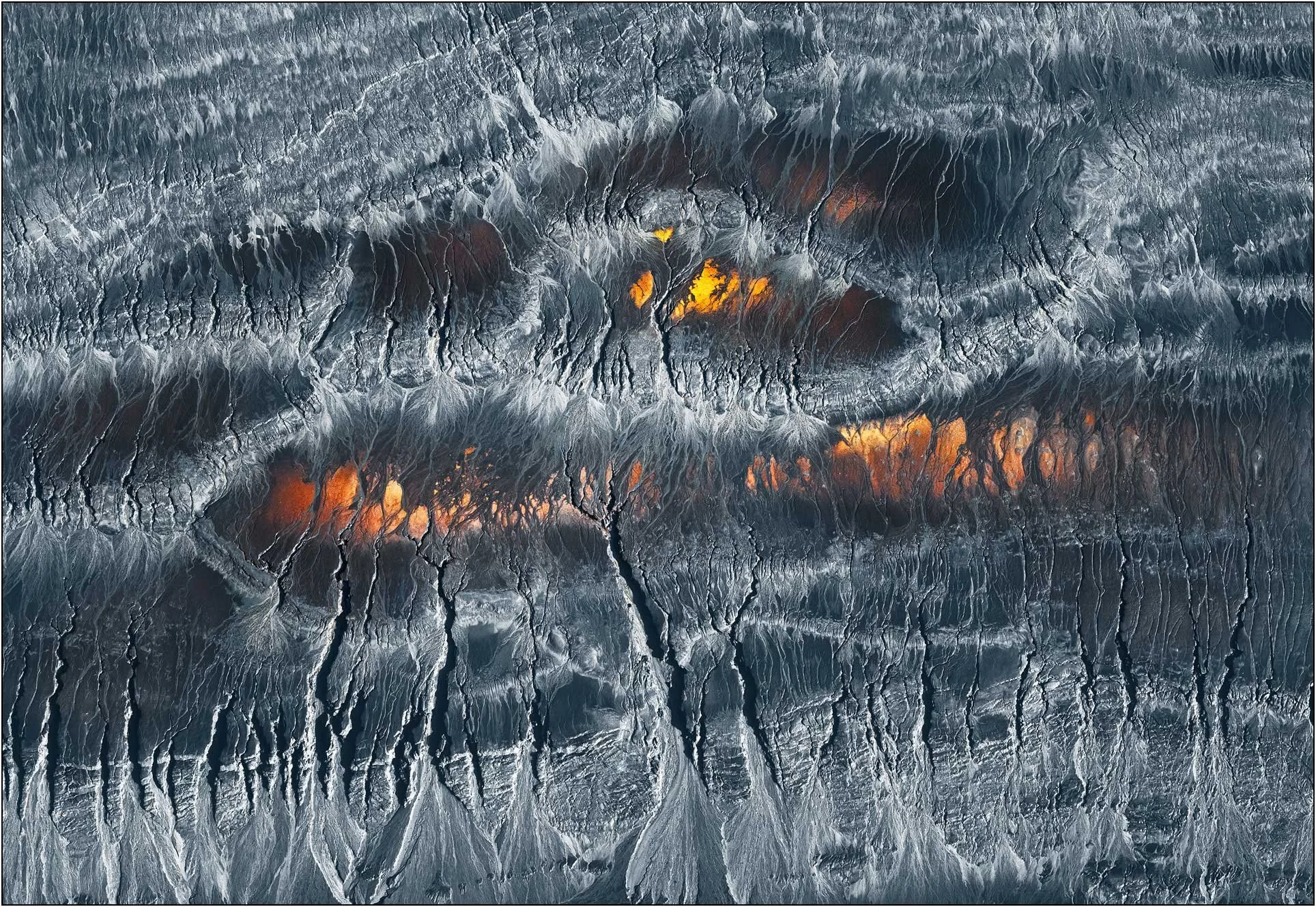 High-contrast aerial abstraction with intricate textures and glowing warm tones beneath layered surfaces