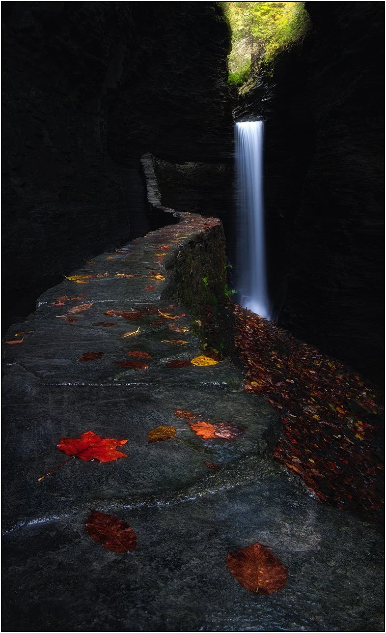 A narrow stone path leading through a canyon toward a vertical waterfall framed by rock walls