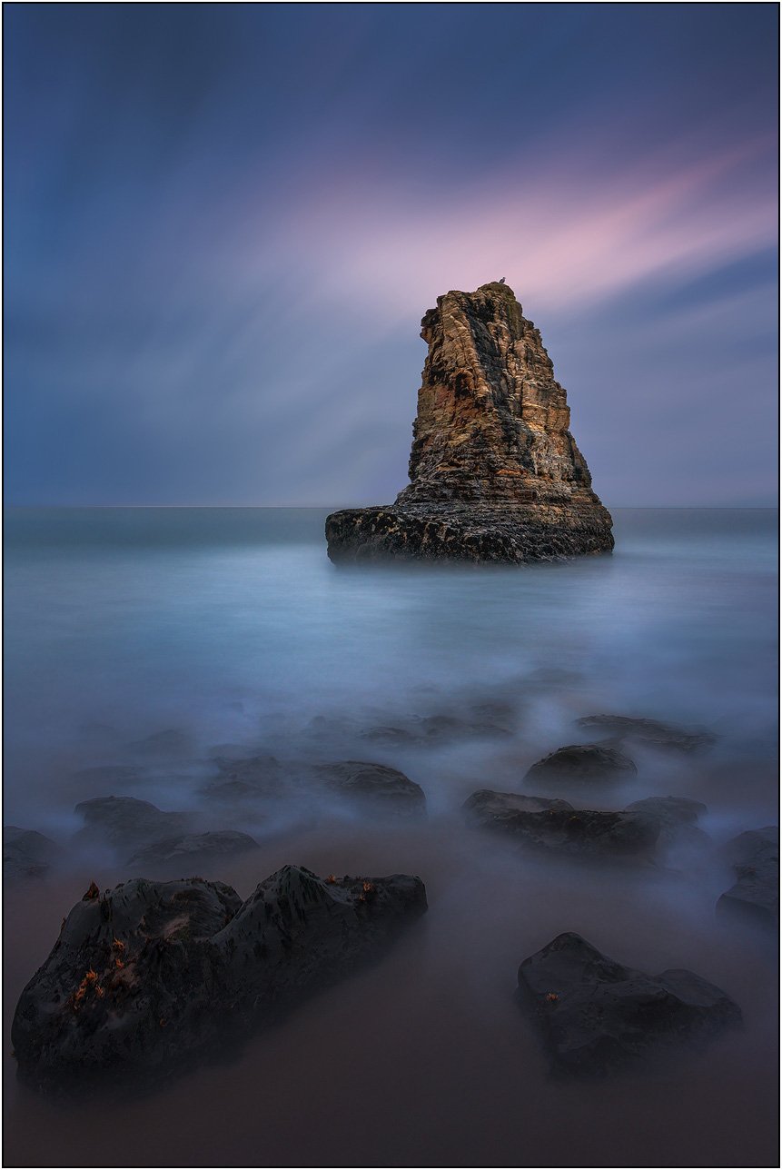 Solitary sea stack rising from calm water at dusk with soft pastel sky