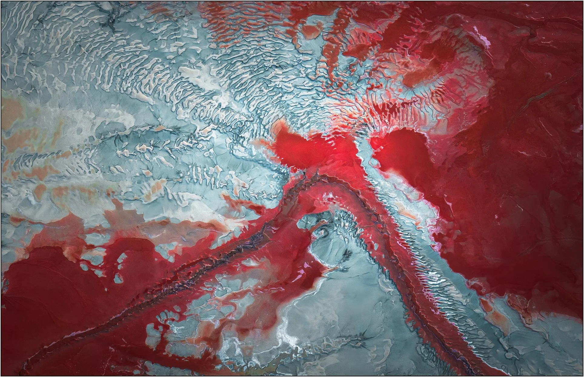 Aerial view of a tailings lake with vivid red mineral patterns spreading across pale textured ground