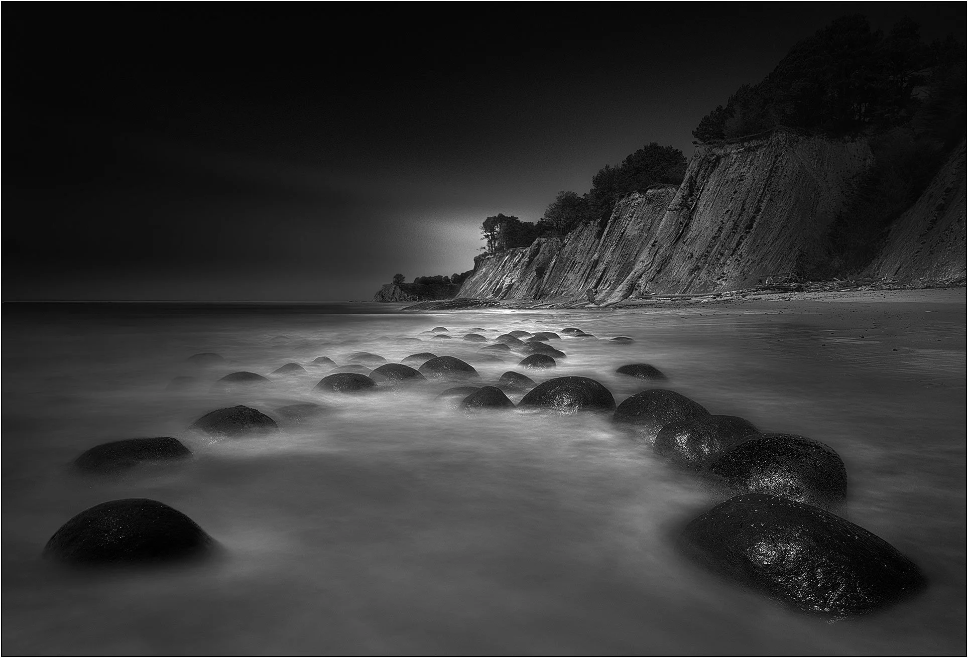 Black and white long exposure of rounded rocks along a shoreline with misty waves and coastal cliffs