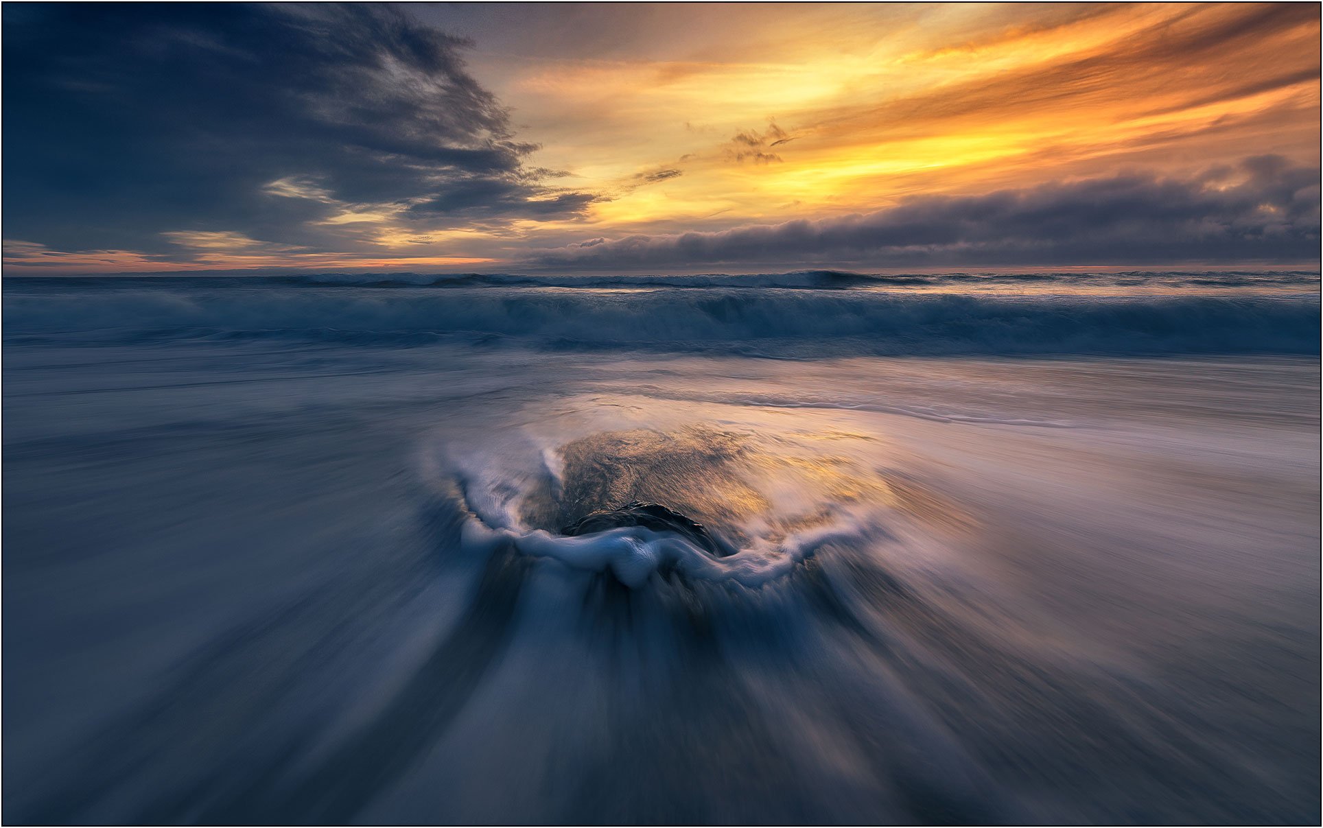 Waves rushing toward a partially submerged rock at sunset, with motion blur and dramatic sky