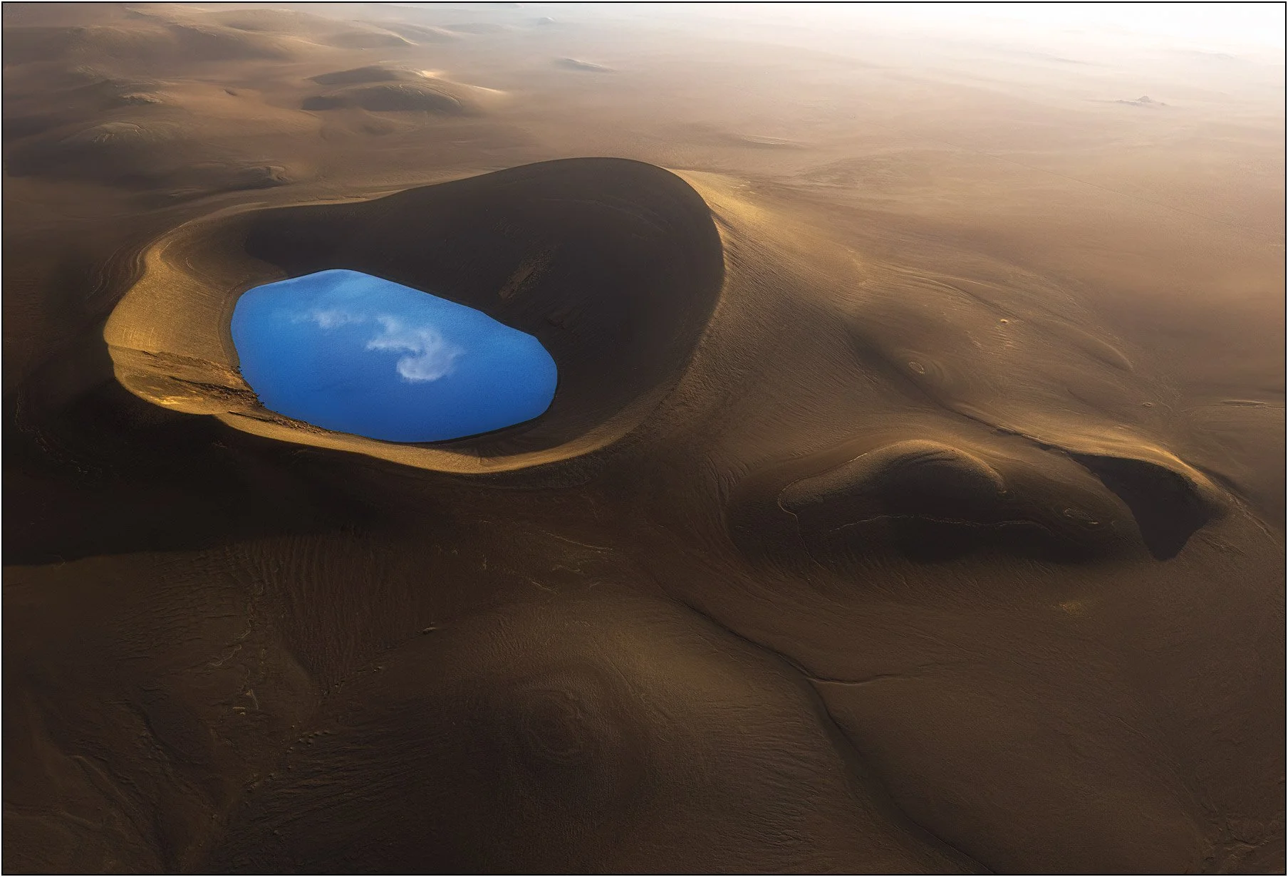 Aerial view of a volcanic crater in Iceland, where a vivid blue lake rests within warm-toned volcanic sand.