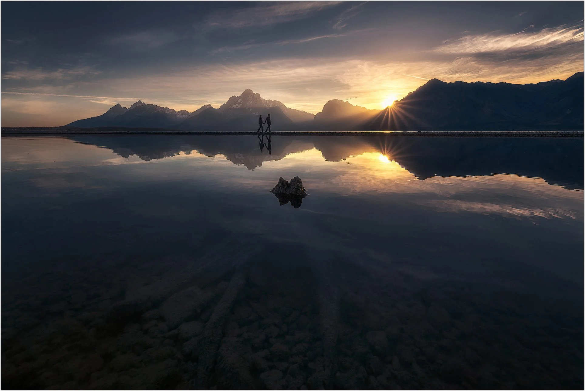 Two people walking along a narrow strip of land at sunrise, with Grand Teton mountain range and reflections in calm water