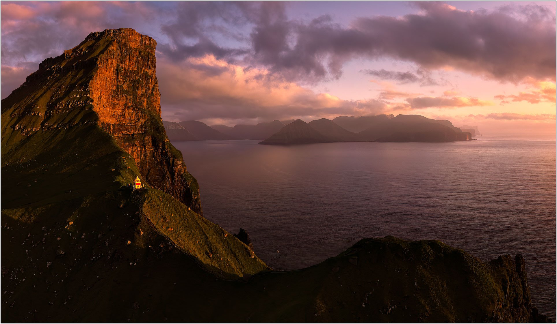 Panoramic aerial view of Kallur lighthouse of Kalsoy, Faroe Islands, with a fjord, mountains, and sunset clouds.