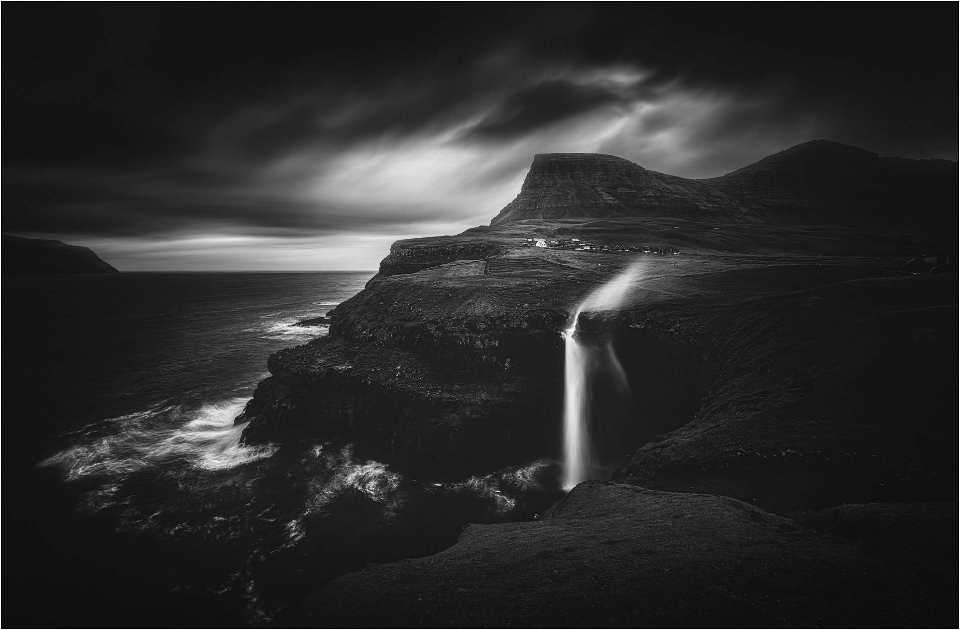 Black and white landscape of Mulafossur waterfall in the Faroe Islands, with a fjord, mountains, long exposure clouds and water blowed by wind.