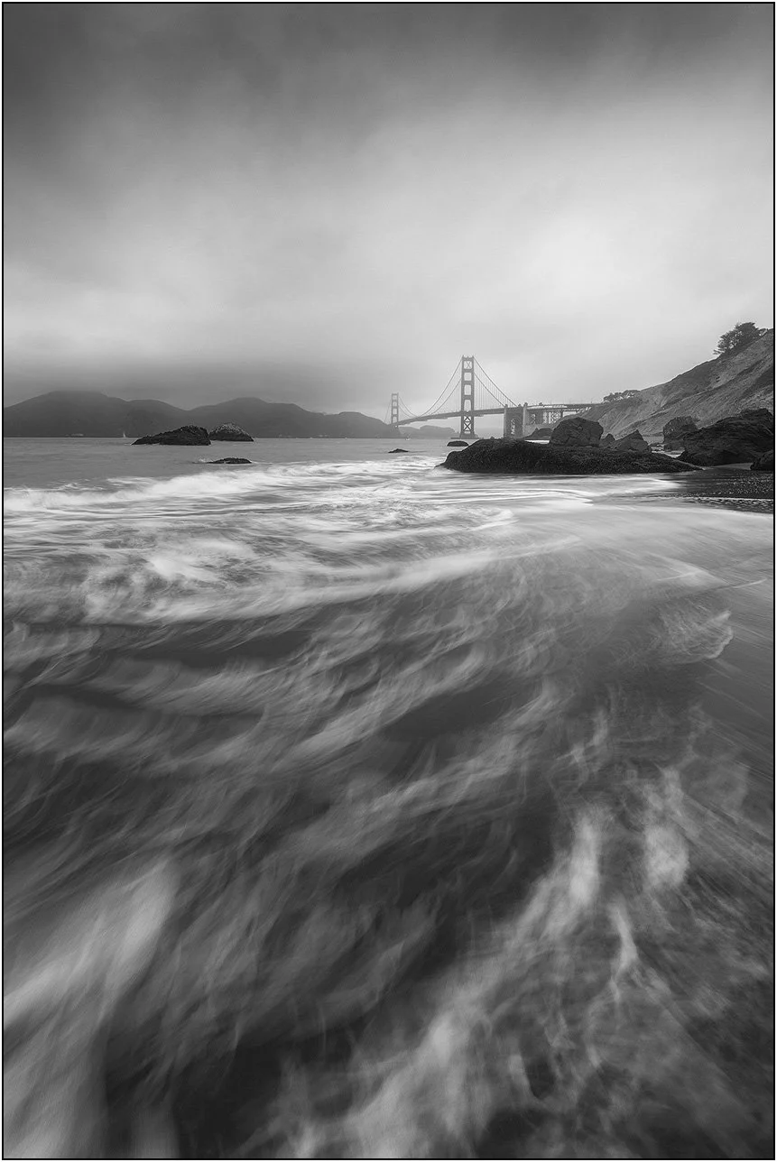 Black and white view of the Golden Gate Bridge from a beach with swirling waves and rocky foreground