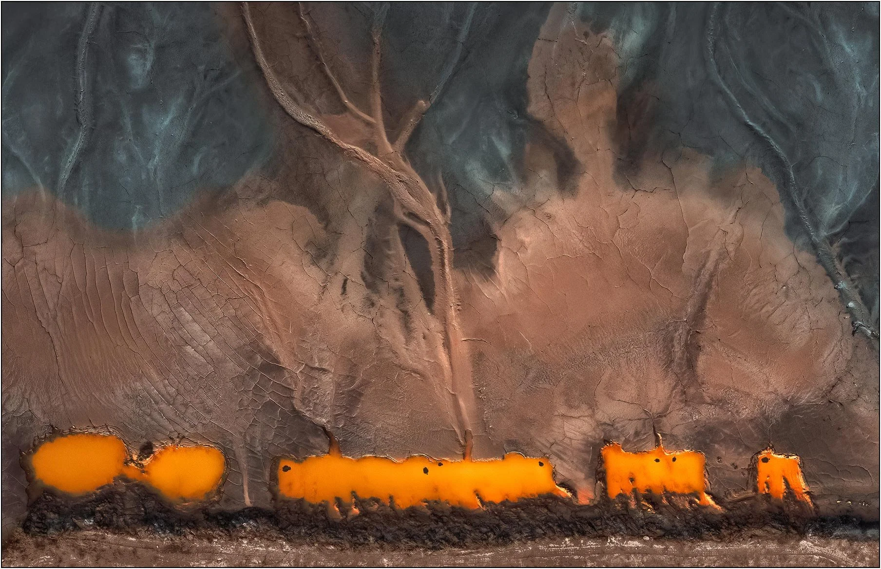 Aerial view of a tailings lake with dark surfaces interrupted by glowing orange mineral bands