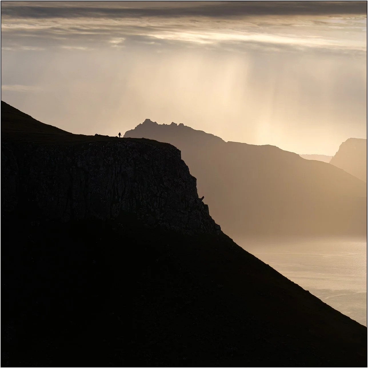 Silhouette of a person standing on a cliff in the Faroe Islands, with dramatic light rays and layered mountains in the background.