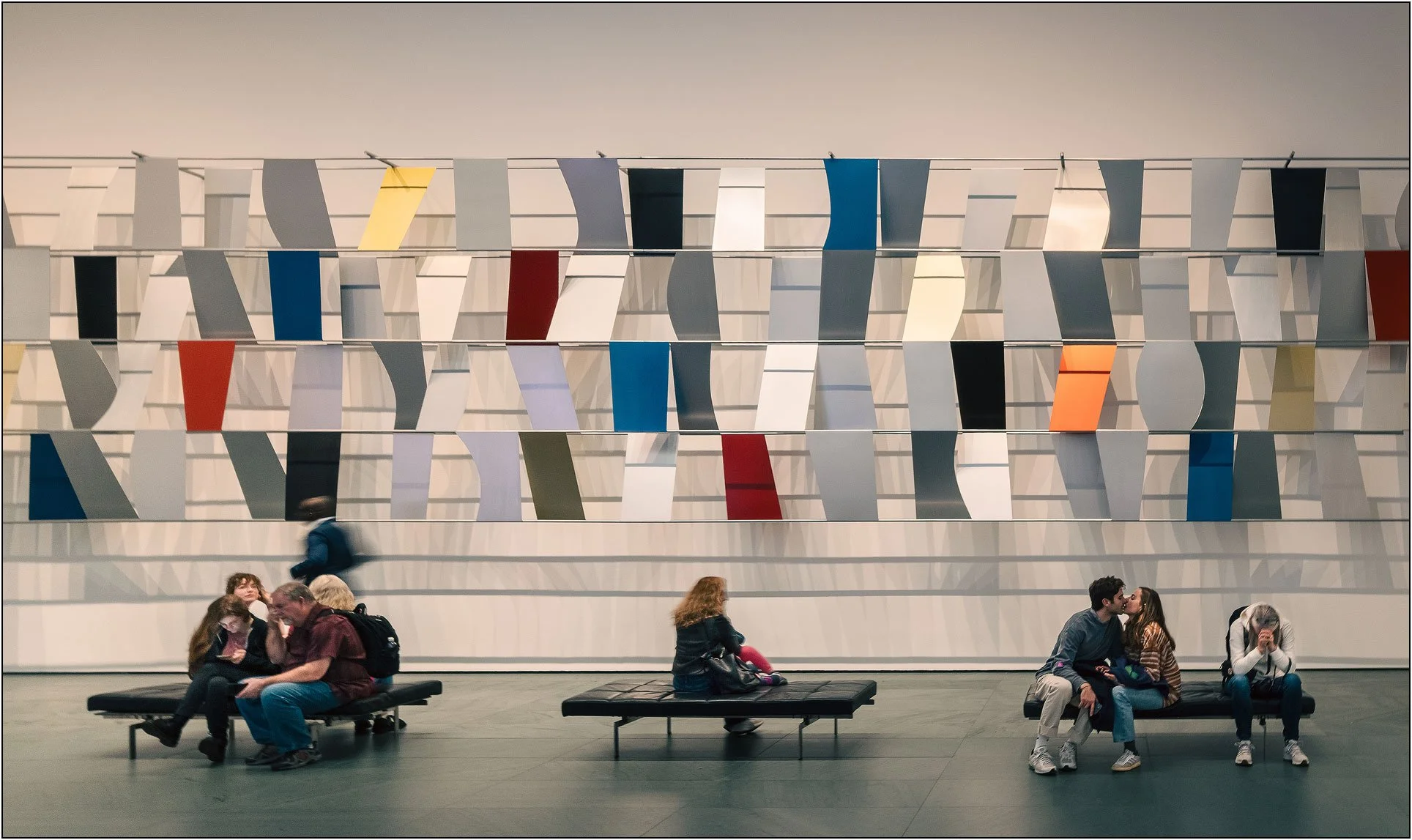 Visitors resting and interacting beneath a rhythmic wall installation of colorful geometric panels in a museum in New York