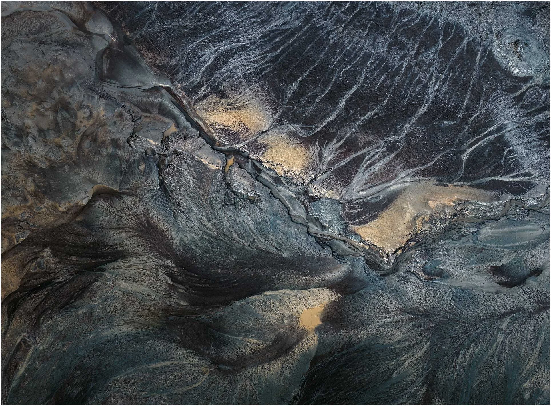 An aerial view of the Qiantang River tidal flats in China, where branching tidal channels and layers of sediment form an intricate natural abstraction. Dark blue-gray textures intersect with warm golden sand, creating patterns reminiscent of river va