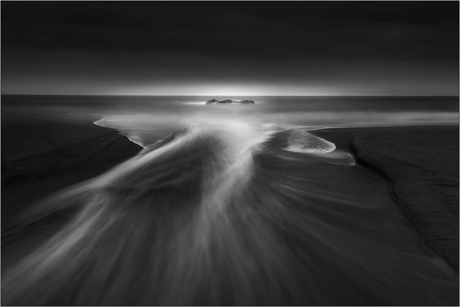 Black and white long exposure of waves flowing across a dark beach toward distant rocks