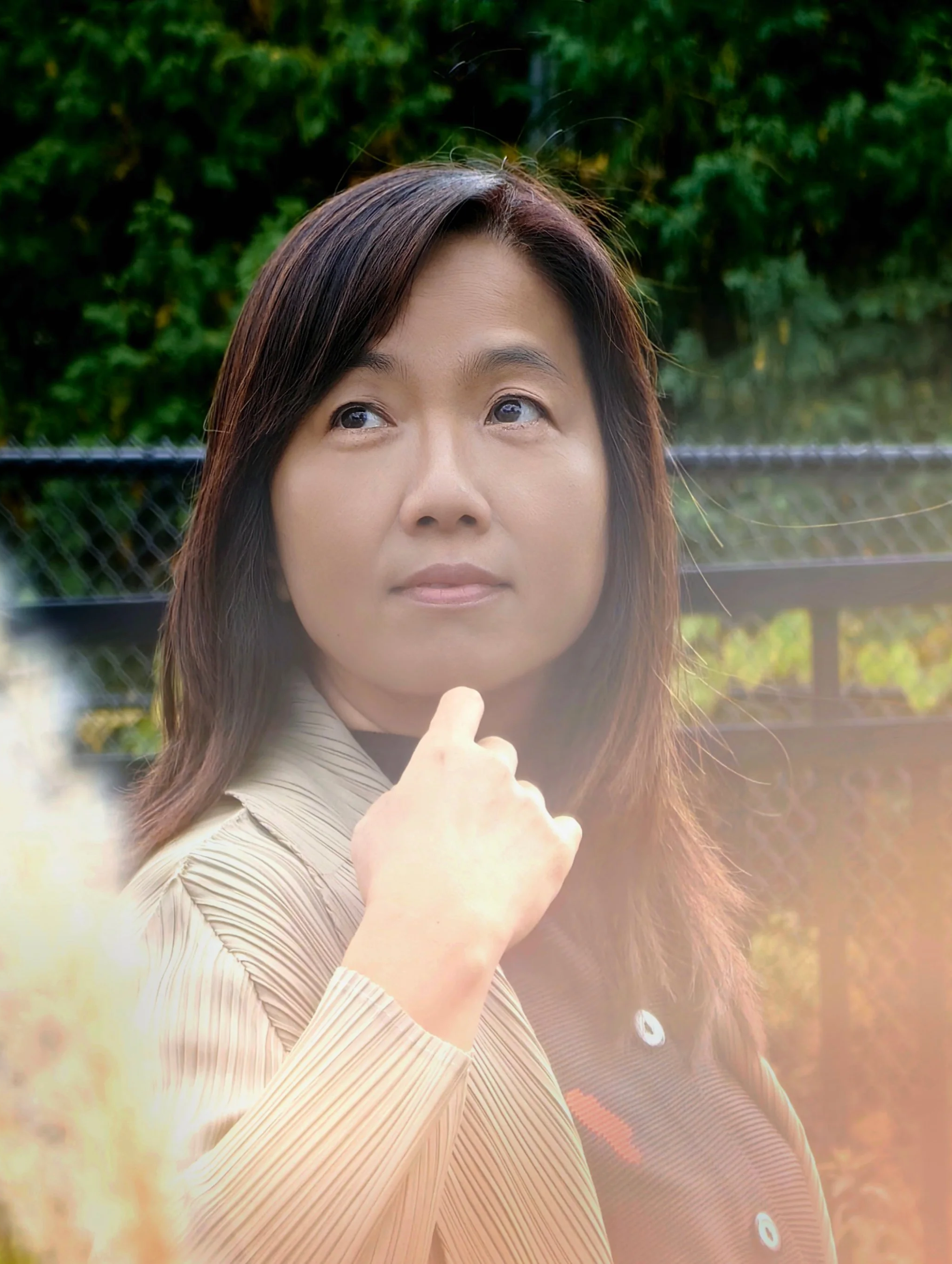 Close-up of a woman with medium-length brown hair, wearing a beige collared shirt, looking upward thoughtfully outdoors with trees and a fence in the background.