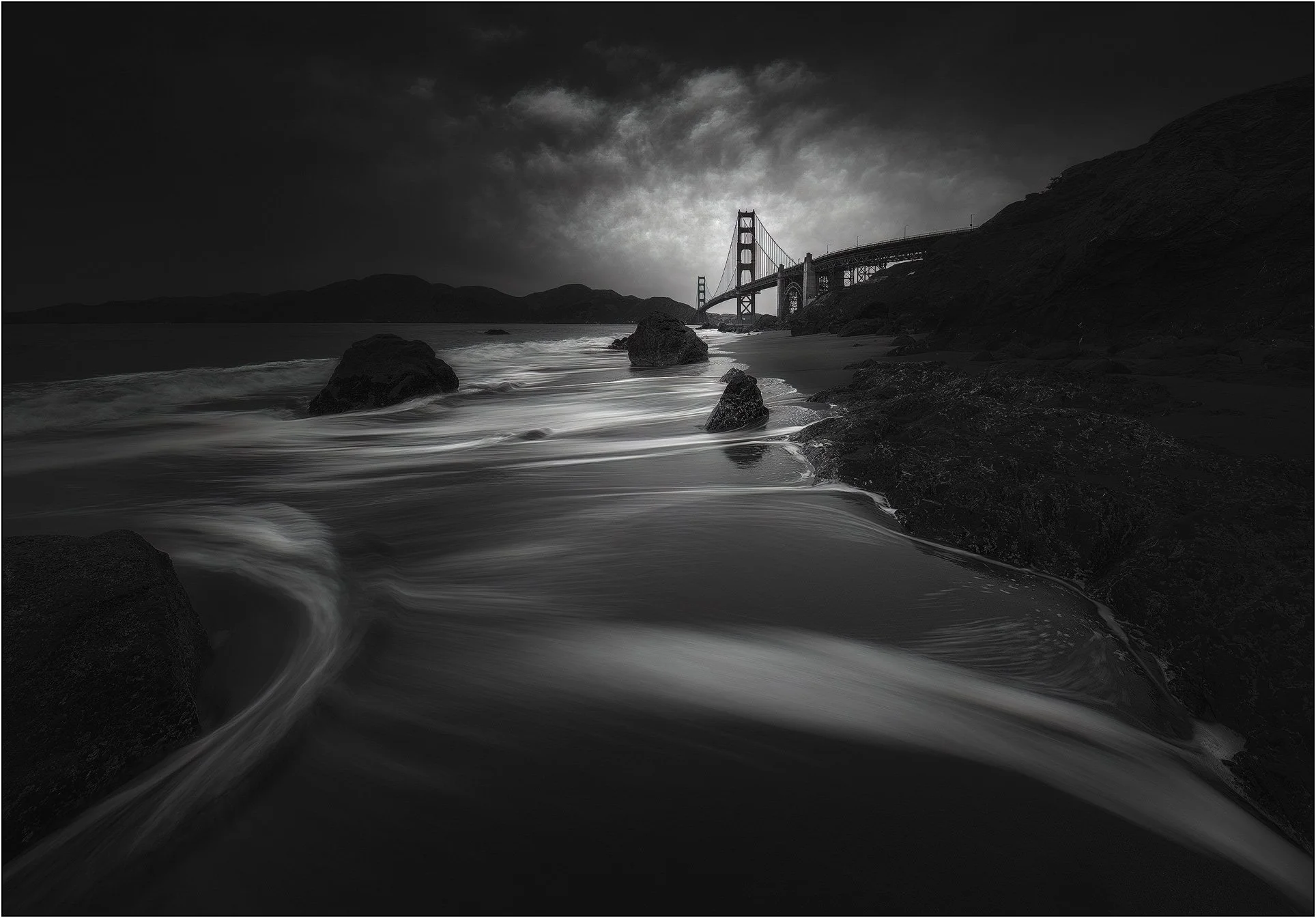 Dark moody black and white seascape with the Golden Gate Bridge in the distance and smooth flowing water