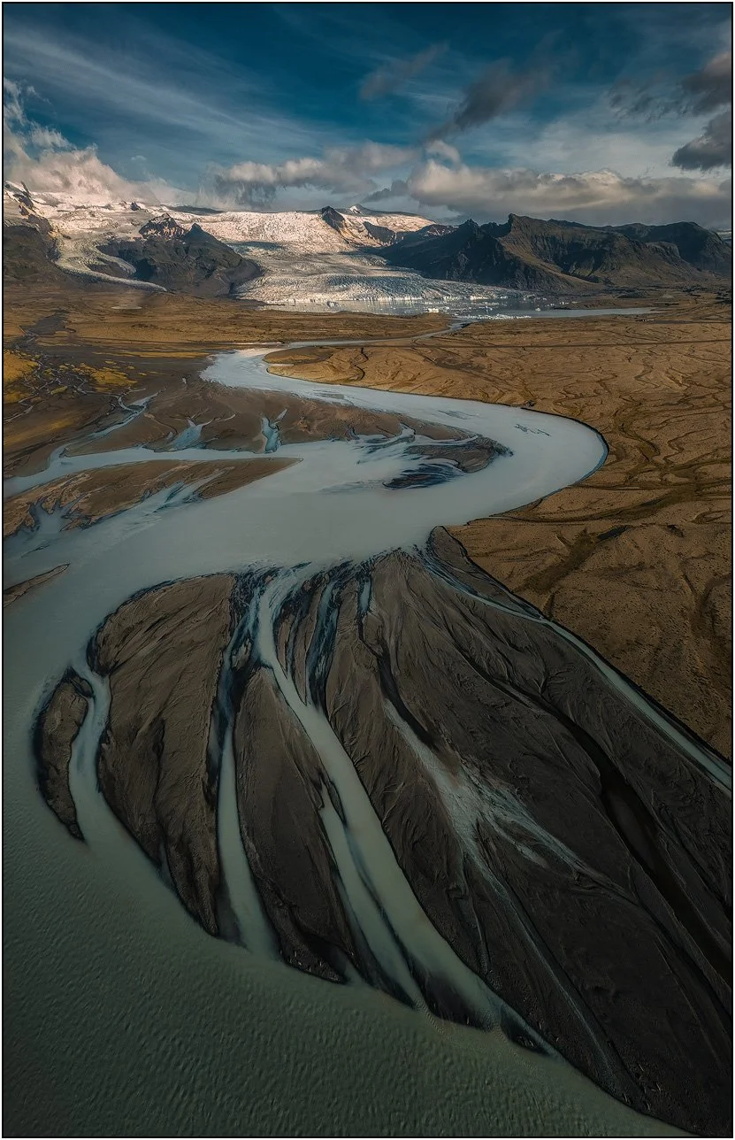 Aerial view of Fjallsjökull, Iceland, with teal colored glacier river flowing over warm toned land, and distant mountains and glacier.