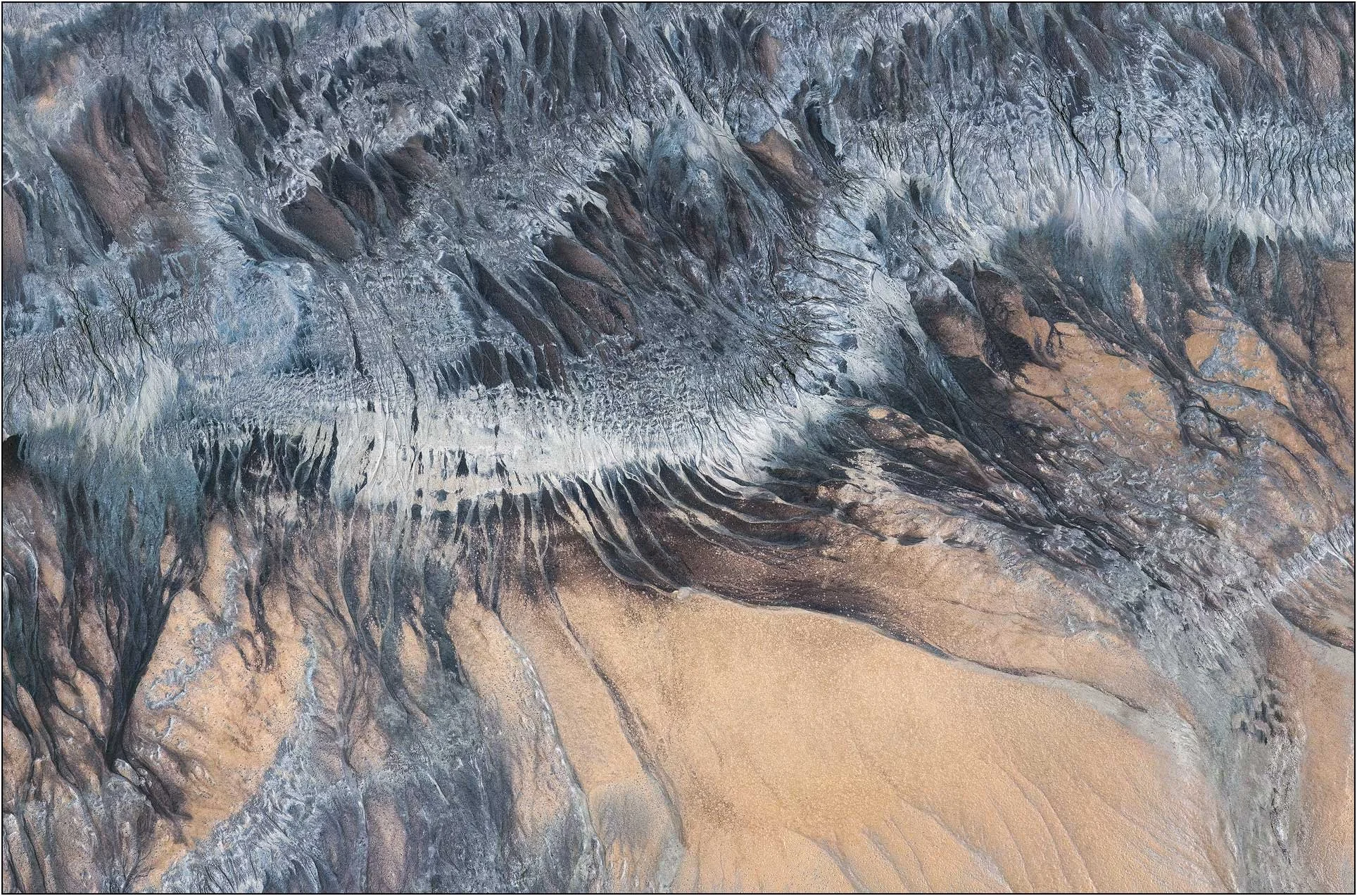 Aerial photography of the Qiantang River tidal flats in China, where flowing water and sediment create intricate, tree-like patterns across the riverbed. Layers of blue-gray, black, and warm sand resemble an imagined mountain landscape, revealing the